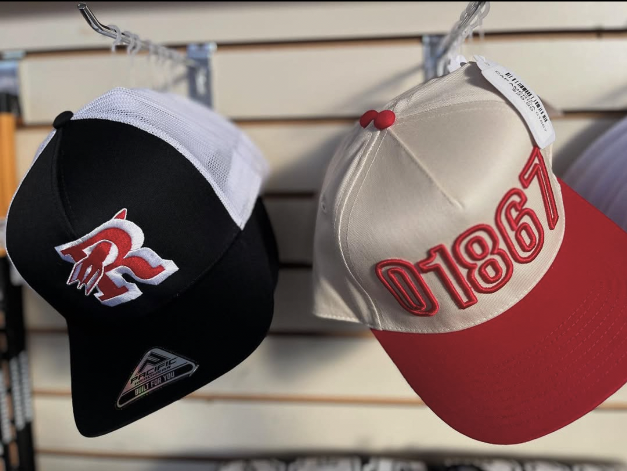 Two baseball caps hanging on a display rack. The cap on the left is black and white with a red and white embroidered logo. The cap on the right is white and red with embroidered red numbers and a white tag.