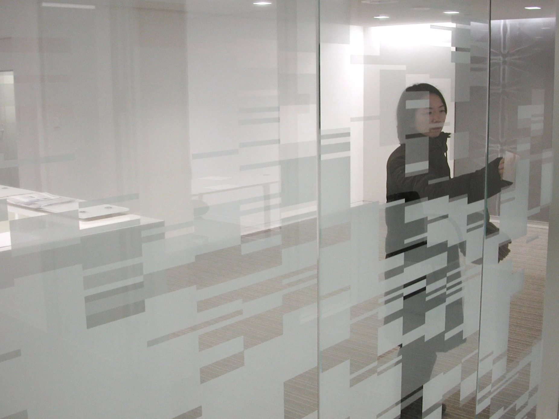 A woman with dark hair, wearing a black coat, standing in an office behind a glass wall with a pattern of frosted rectangles, holding a smartphone in her left hand.