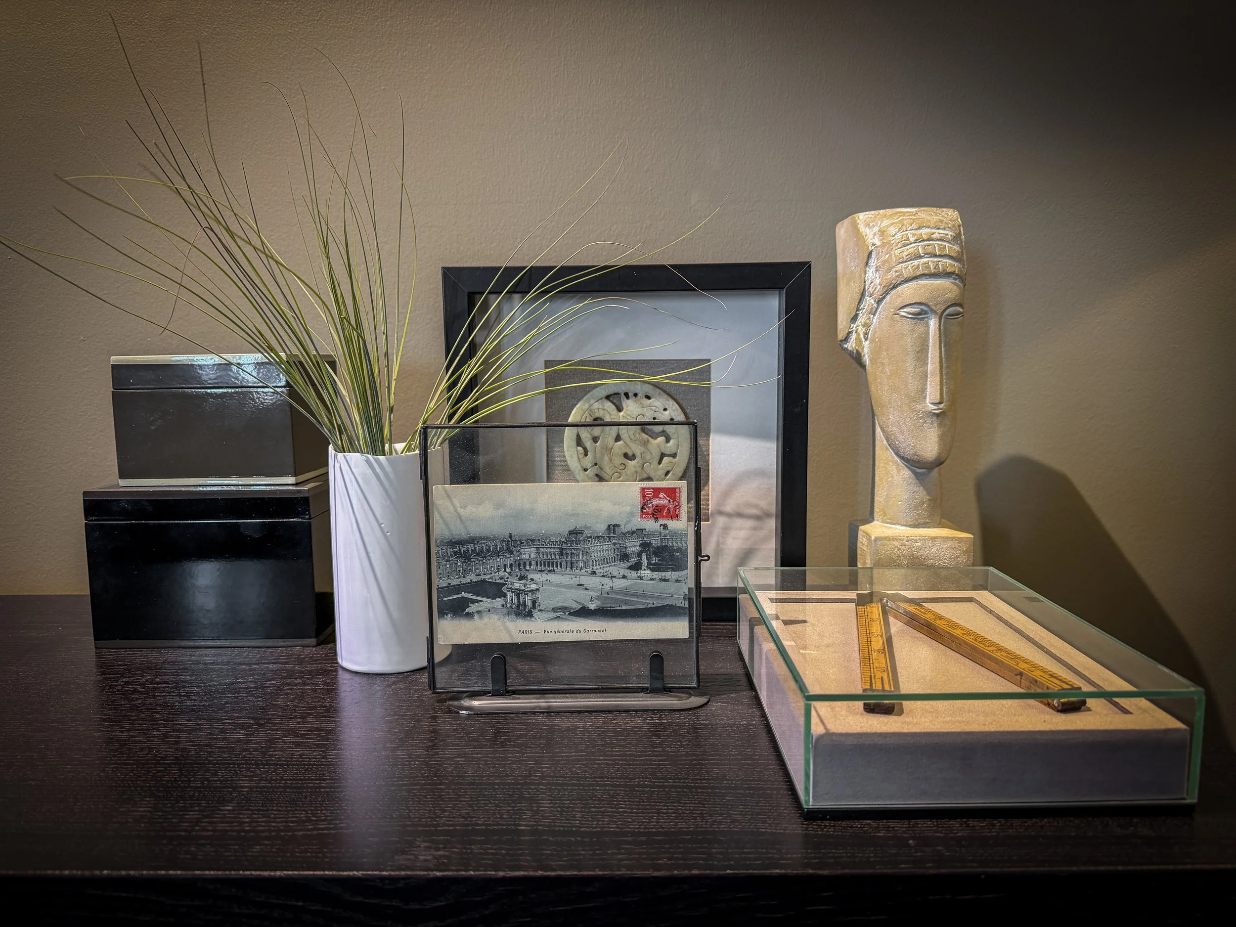 Decorative objects on a dark wooden surface including a black and white striped box, a tall white vase with grass, a framed black and white artwork, a stone sculpture of a human face, and a glass display case with rulers inside.