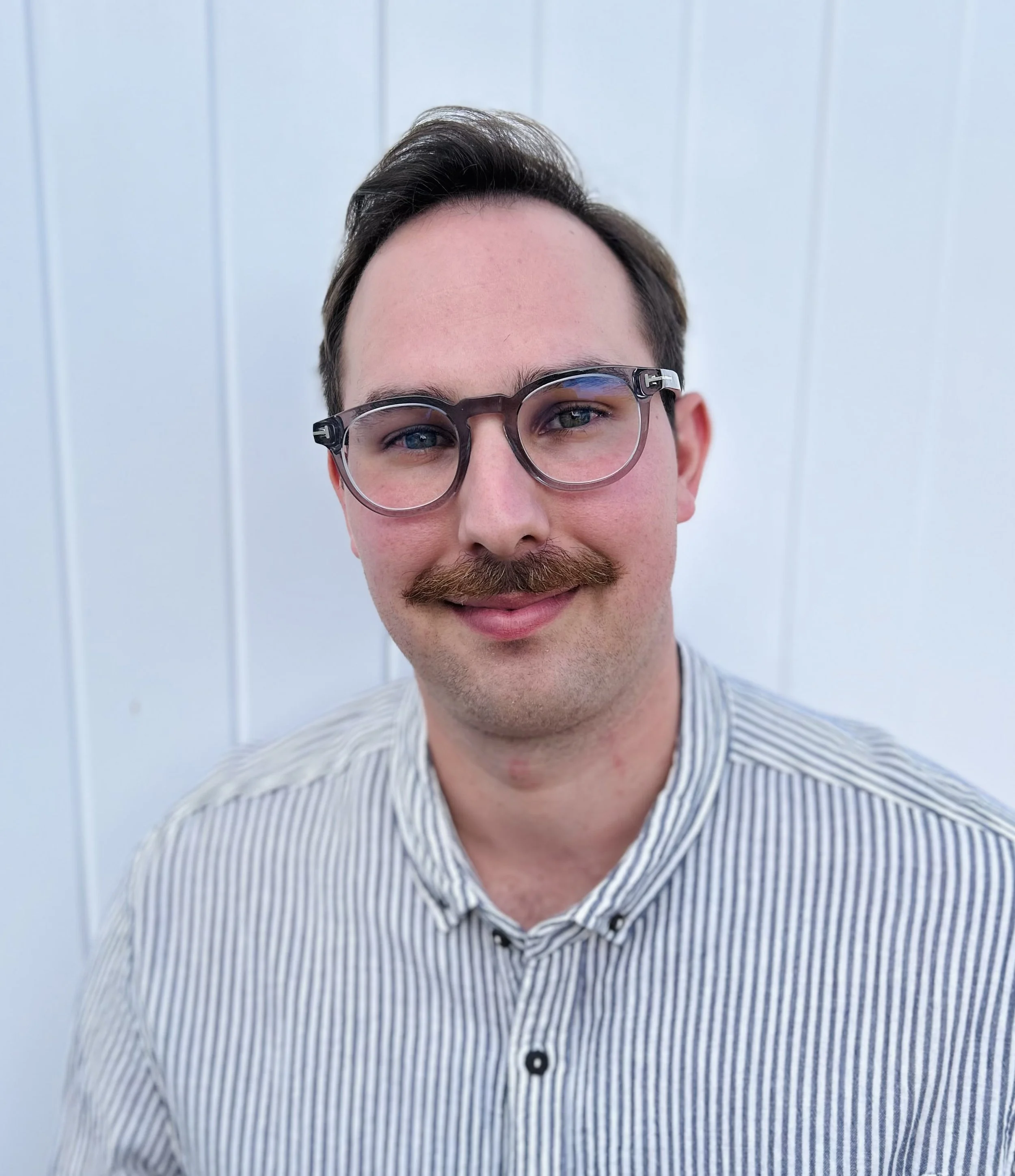 A man with glasses, a mustache, and a striped shirt smiling in front of a white background.