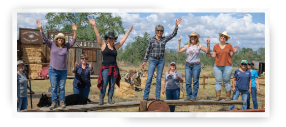 A group of women standing on a wooden fence waving to the camera in a ranch environment on a sunny day.