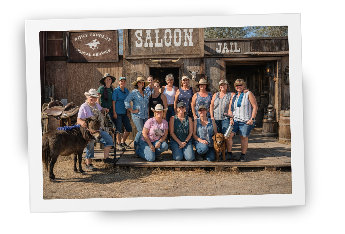 Group of women gathered in front of a weathered wooden saloon building with a miniature donkey and dog, standing and kneeling together on the ranch.