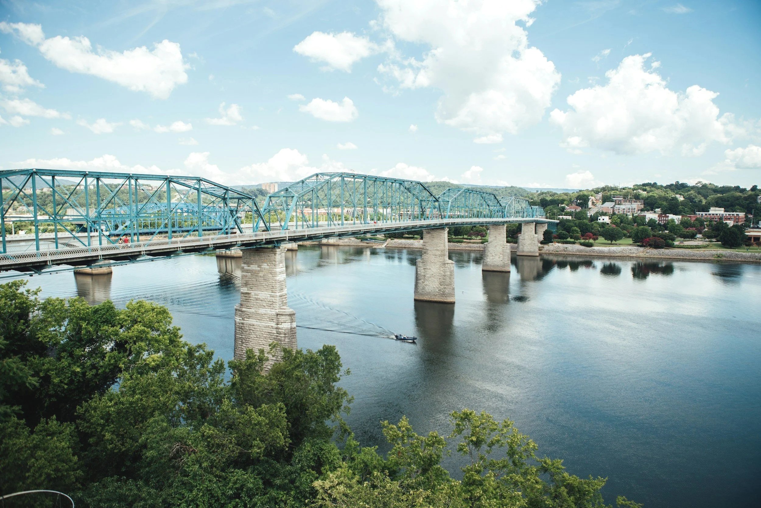 View of a blue truss bridge crossing a wide river, with a small boat sailing underneath, surrounded by green trees and a partly cloudy sky.