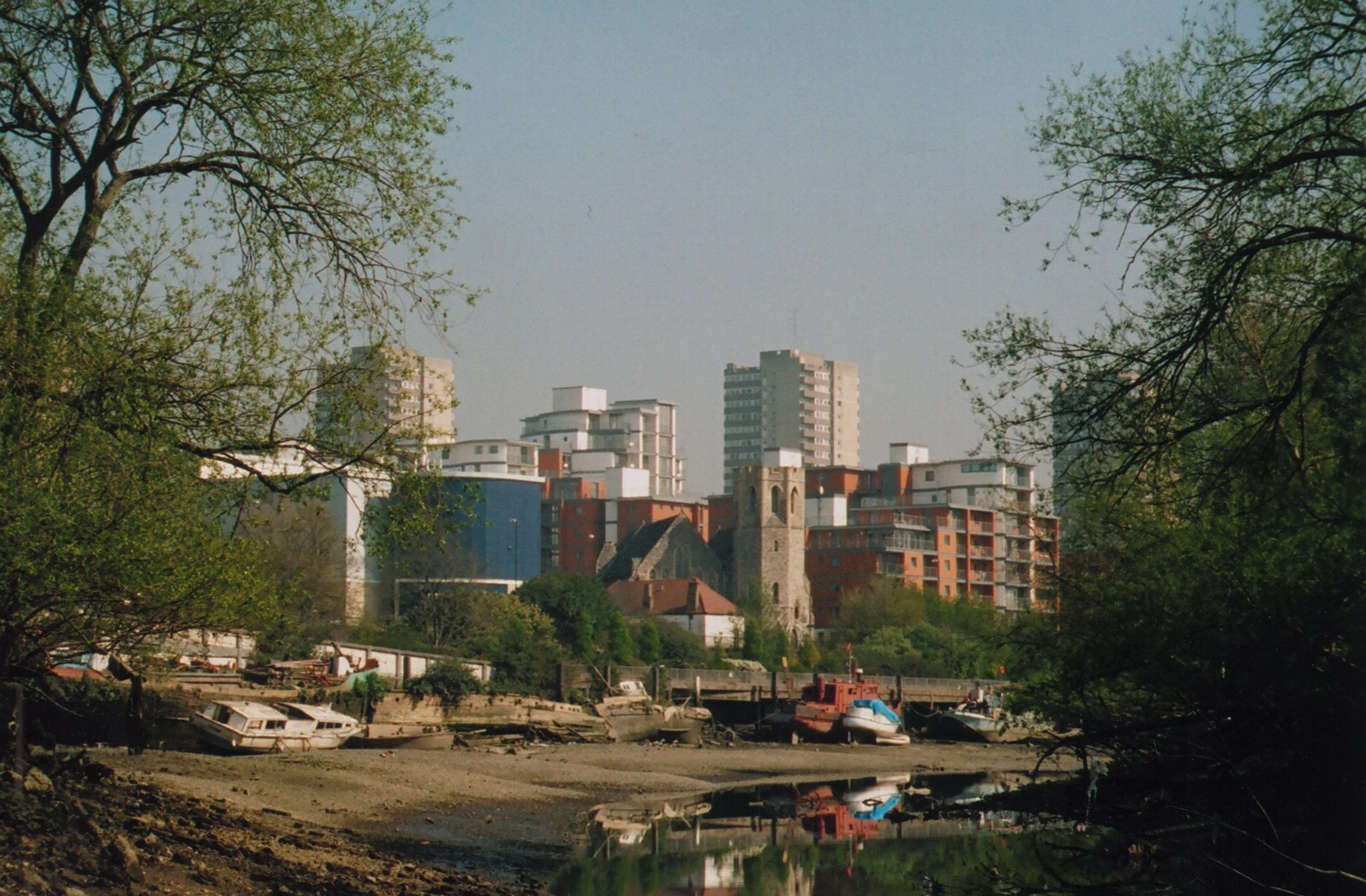 Urban industrial skyline of Brentford framed by trees with the river Thames at low tide showing foreshore