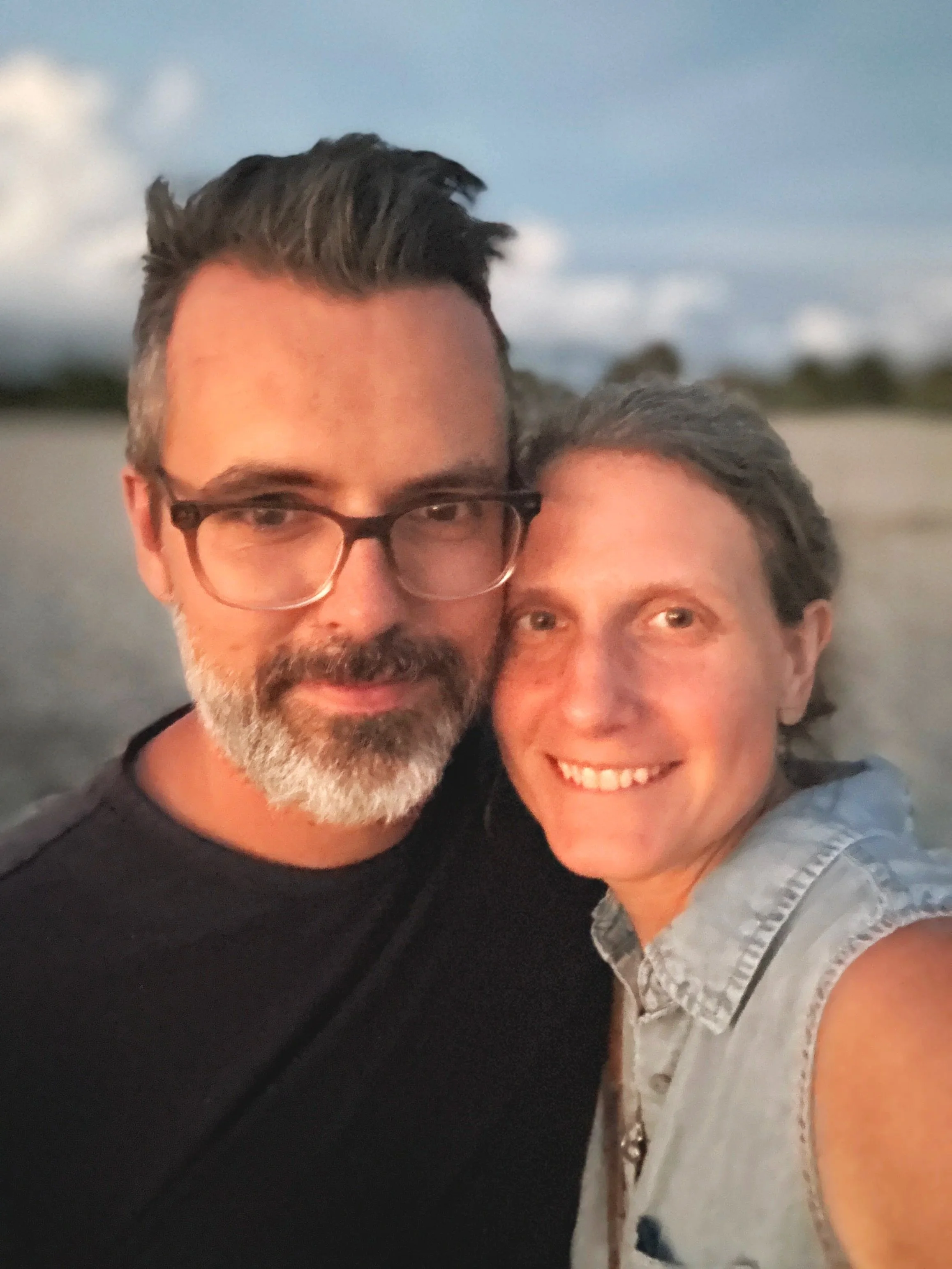 A smiling man with glasses and a beard taking a selfie with a woman on a beach at sunset.