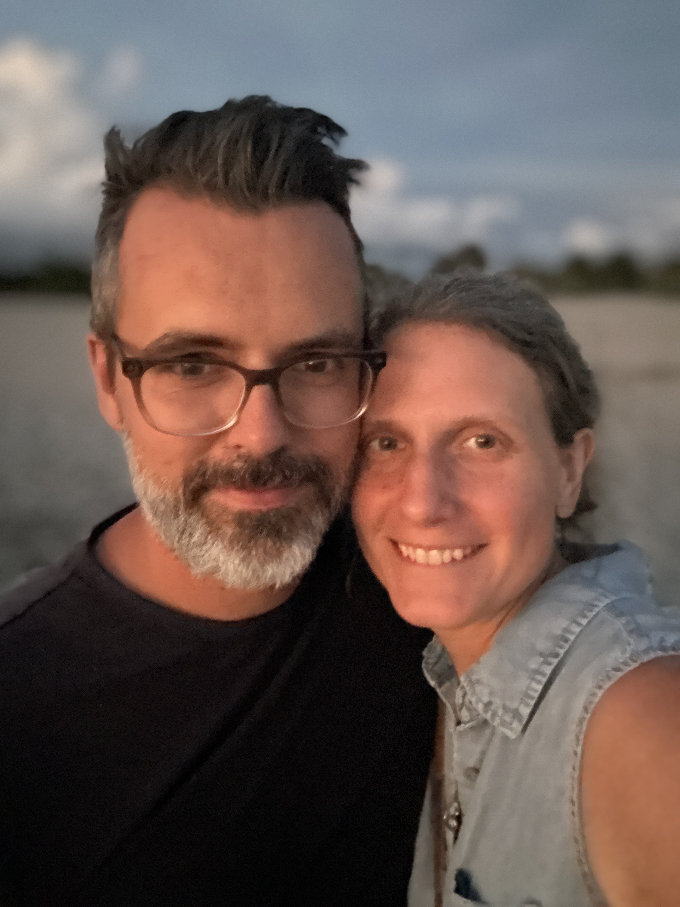 A couple taking a close-up selfie outdoors at sunset or dusk, with a blurred beach and sky in the background.