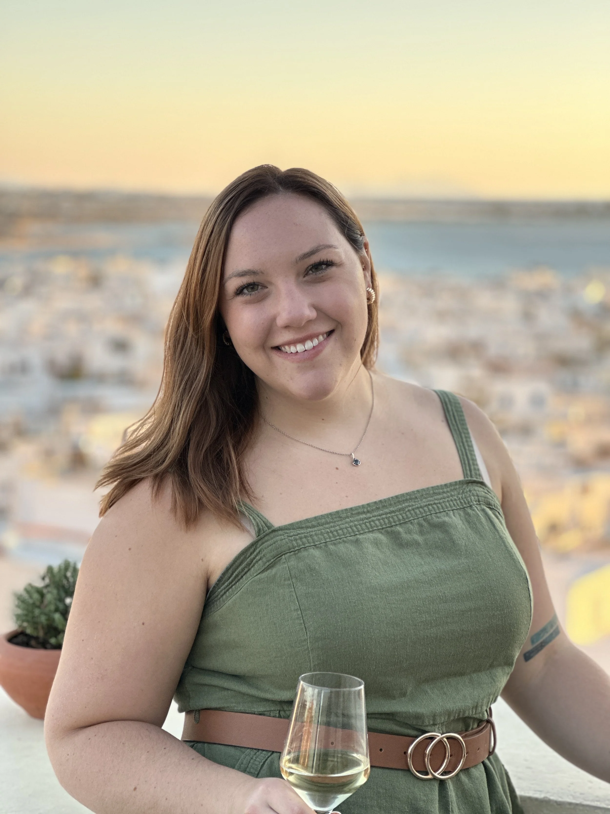 A woman with brown hair smiling at the camera while holding a glass of white wine, with a city skyline and a body of water in the background during sunset.