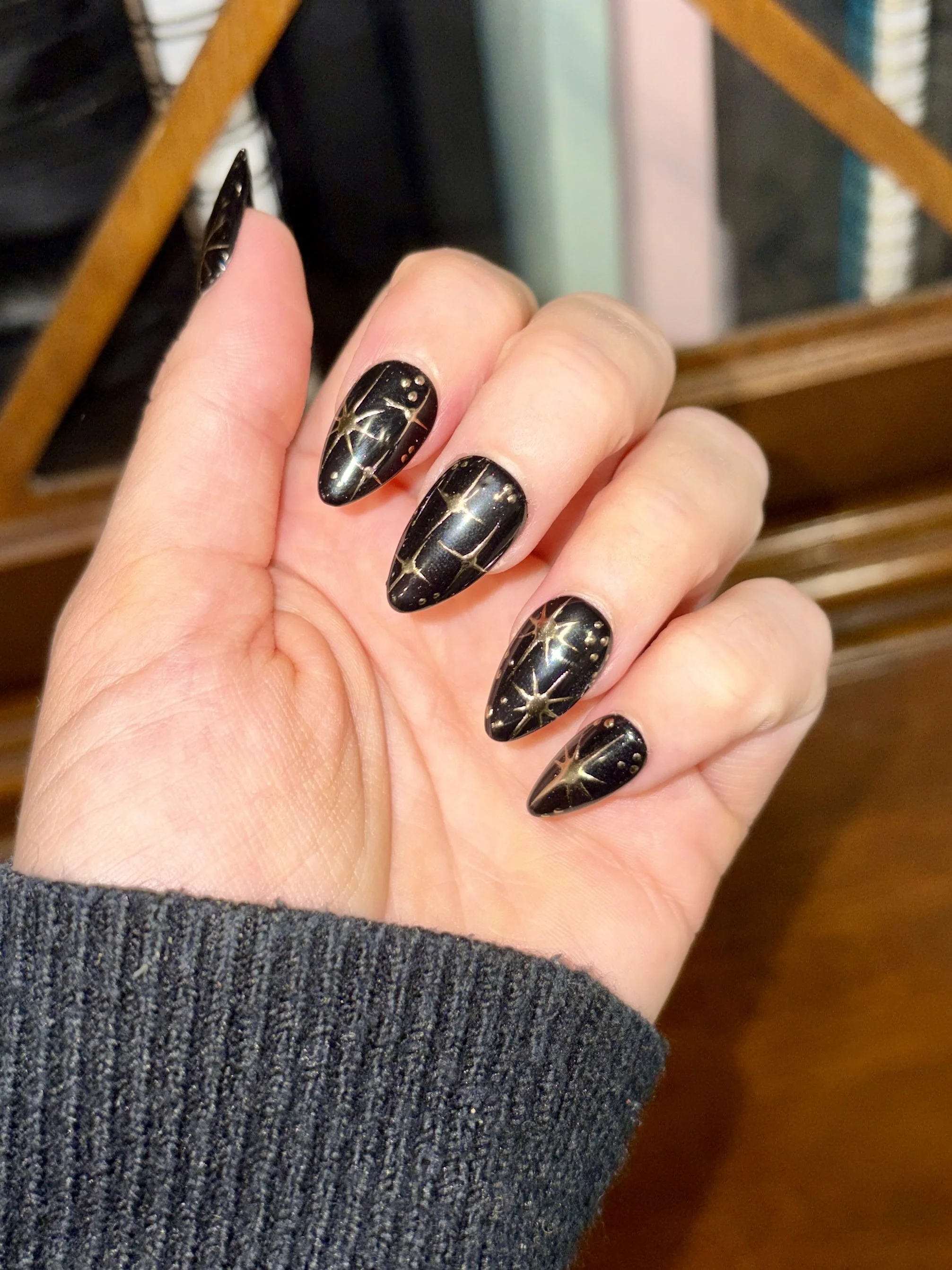 Hand with black, glossy, pointed nails featuring gold star and dot designs, wearing a gray sweater.