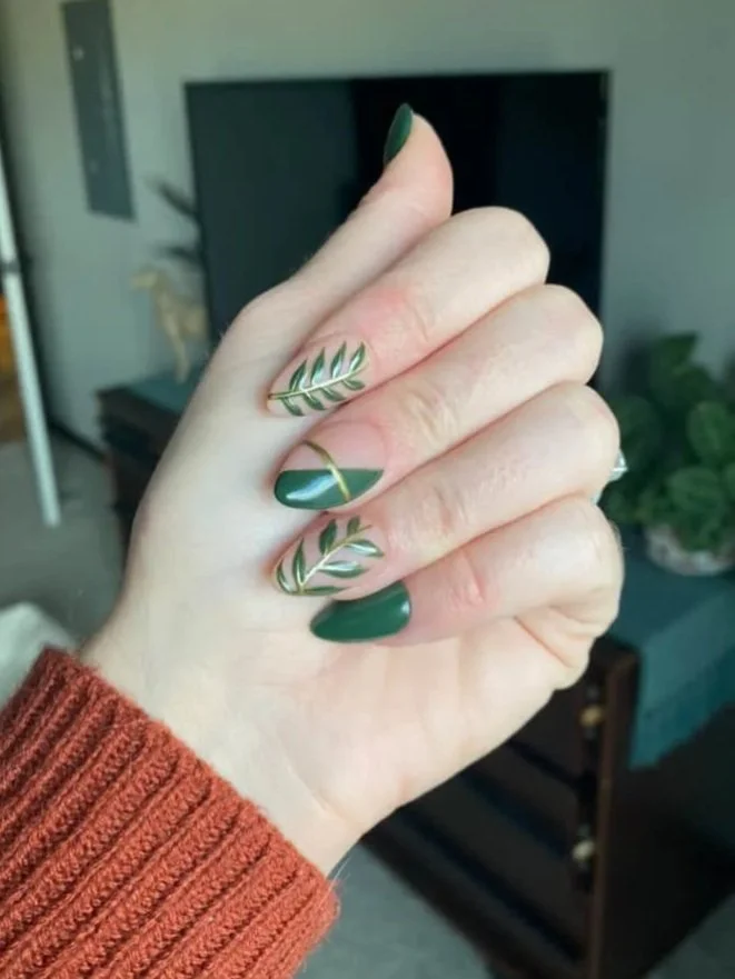 Hand with manicured nails featuring dark green and gold leaf designs, wearing an orange sweater, with a blurred living room in the background.