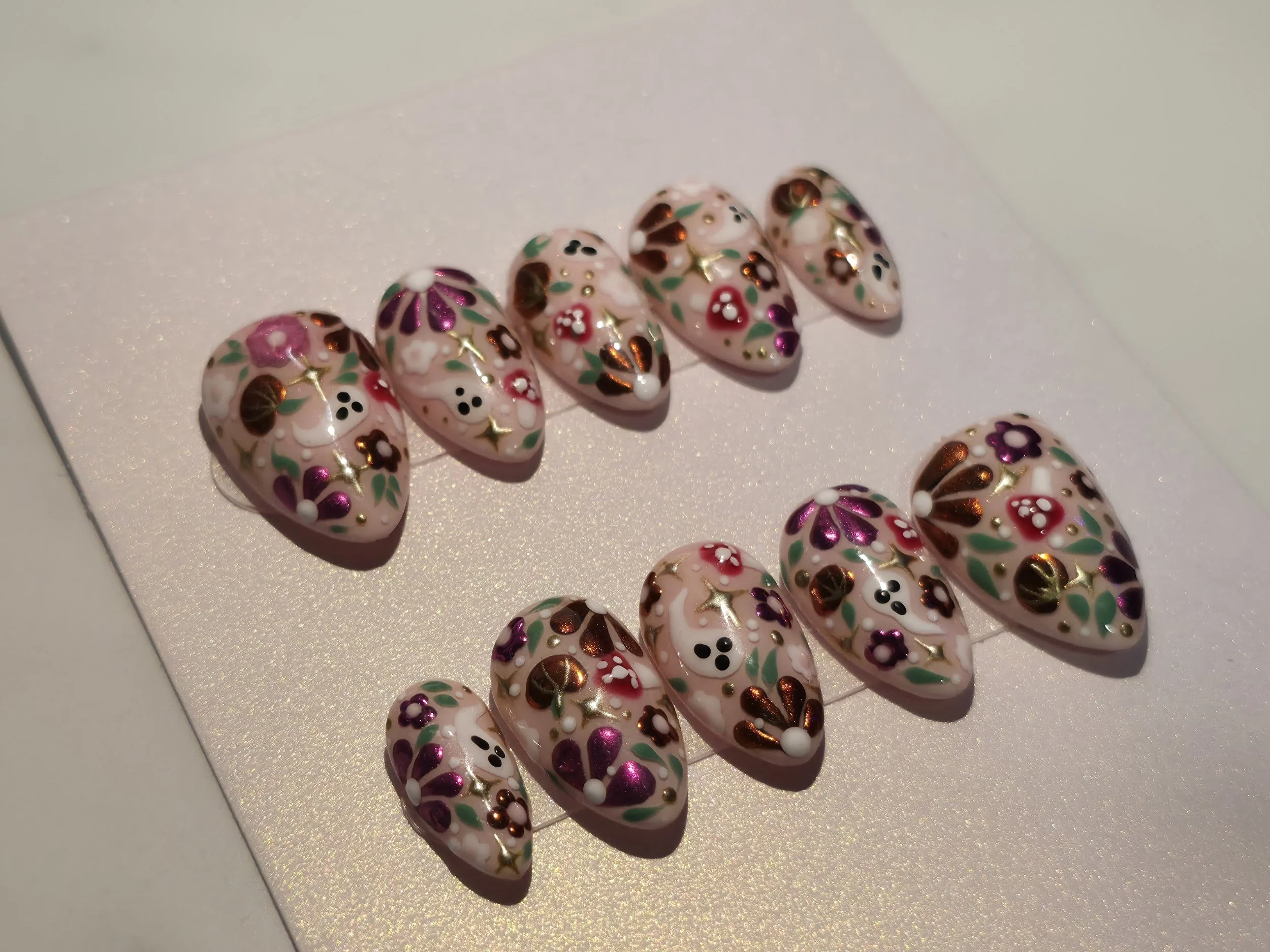 Set of decorative, floral nail art designs on oval-shaped nails with a pink background and gold, white, purple, and green accents.