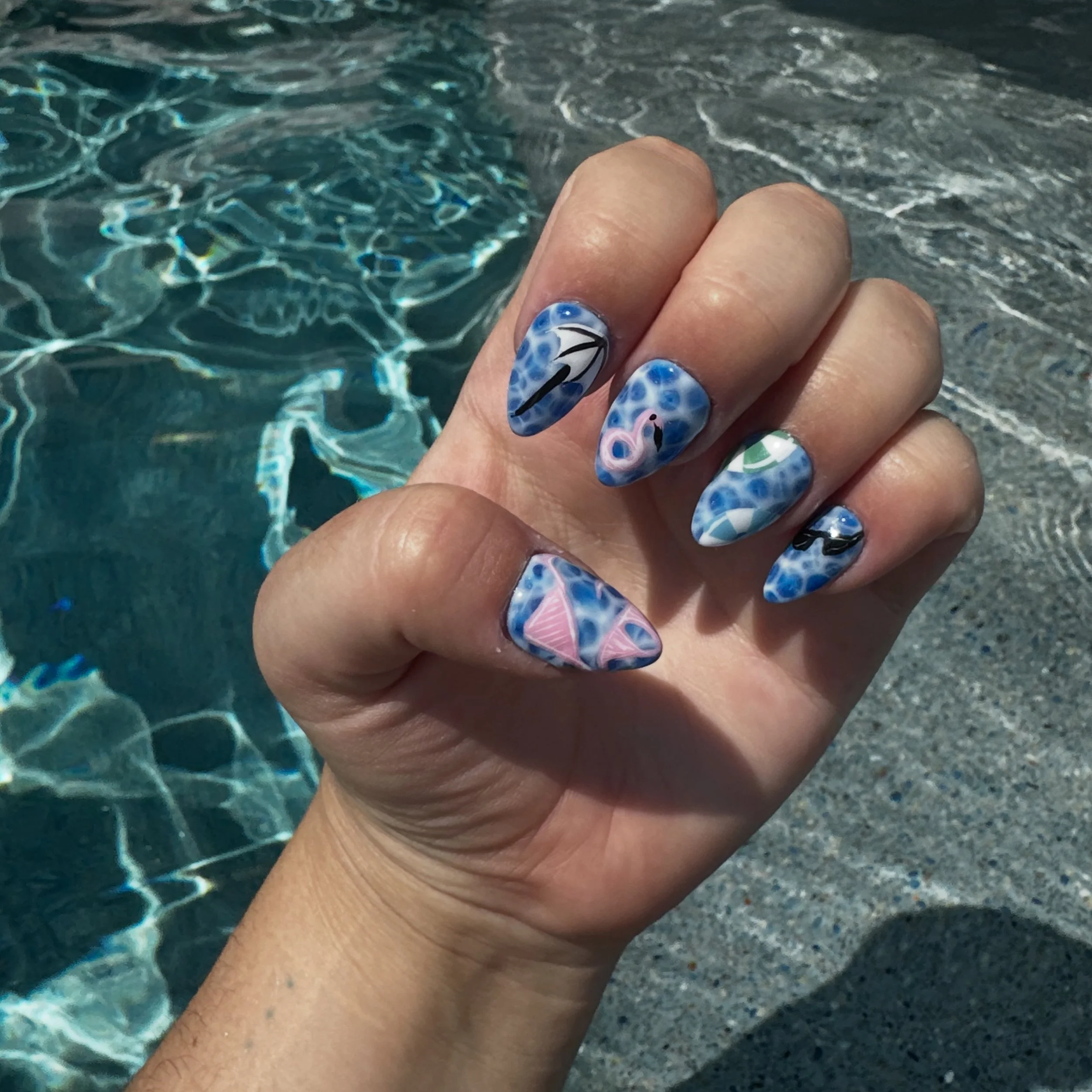 Hand showing decorated nails with abstract colorful designs, near a swimming pool with rippling water.