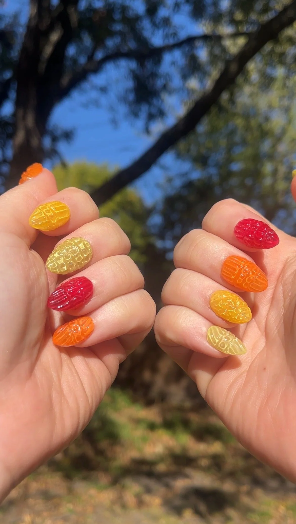 Hands with colorful gummy bear nail art, held up outdoors with trees and blue sky in the background.