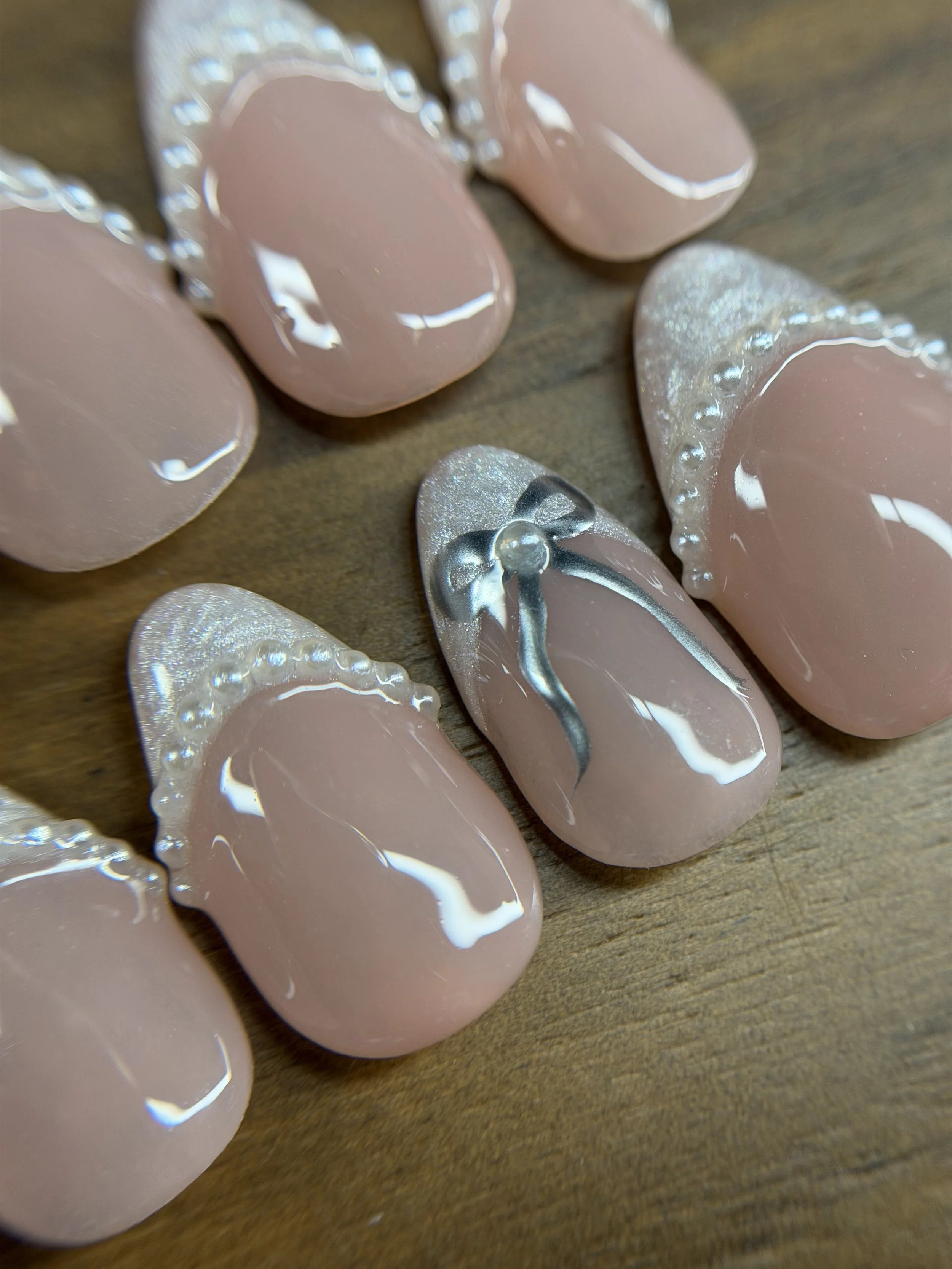 Close-up of decorative pink and silver artificial nails with pearl and bead embellishments, arranged on a wooden surface.