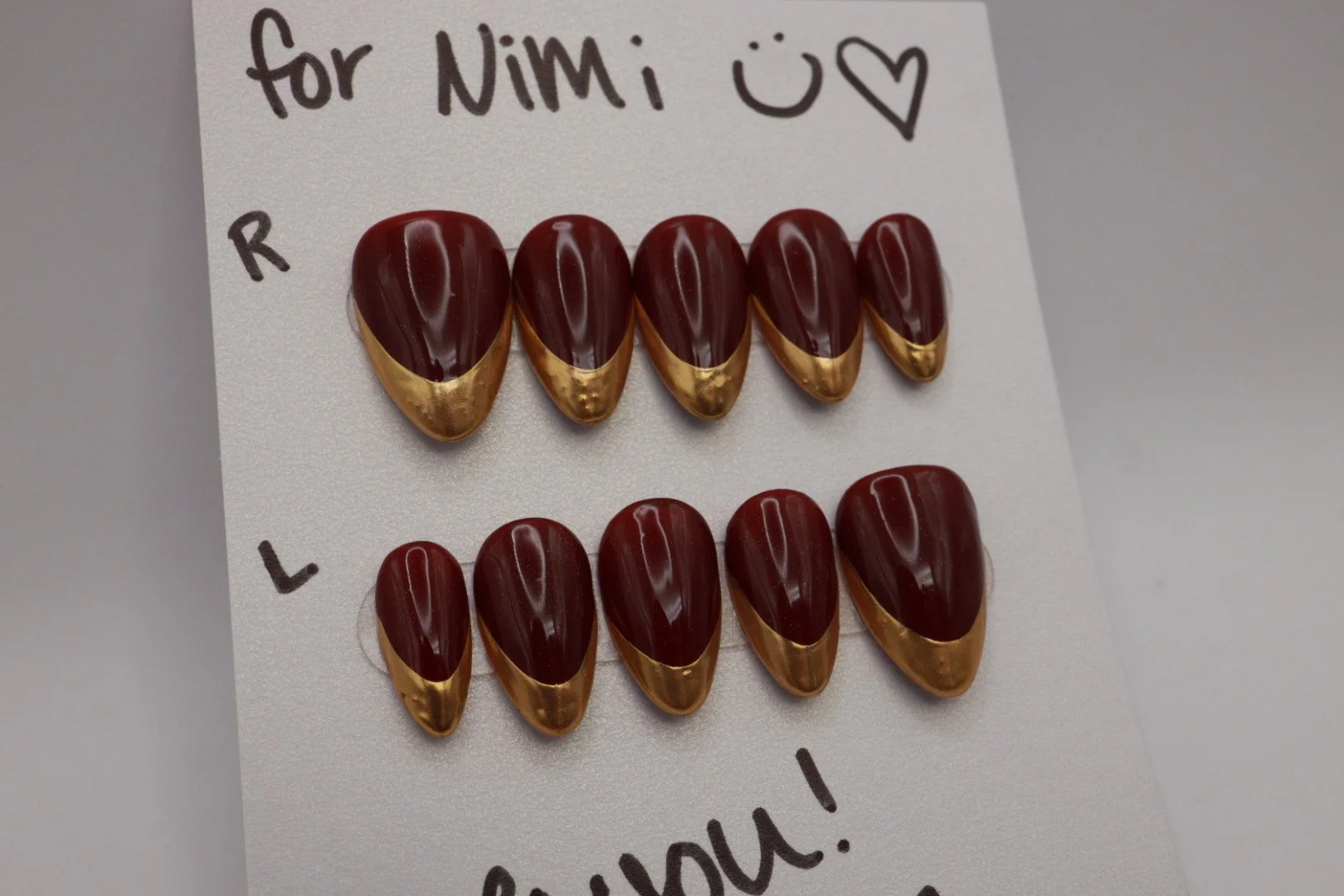 Two rows of decorated almonds on a white card with handwritten notes. The top row labeled 'R' has five almonds with gold and dark chocolate coating, and the bottom row labeled 'L' has five almonds similarly decorated. The card says 'for Nimi' at the 