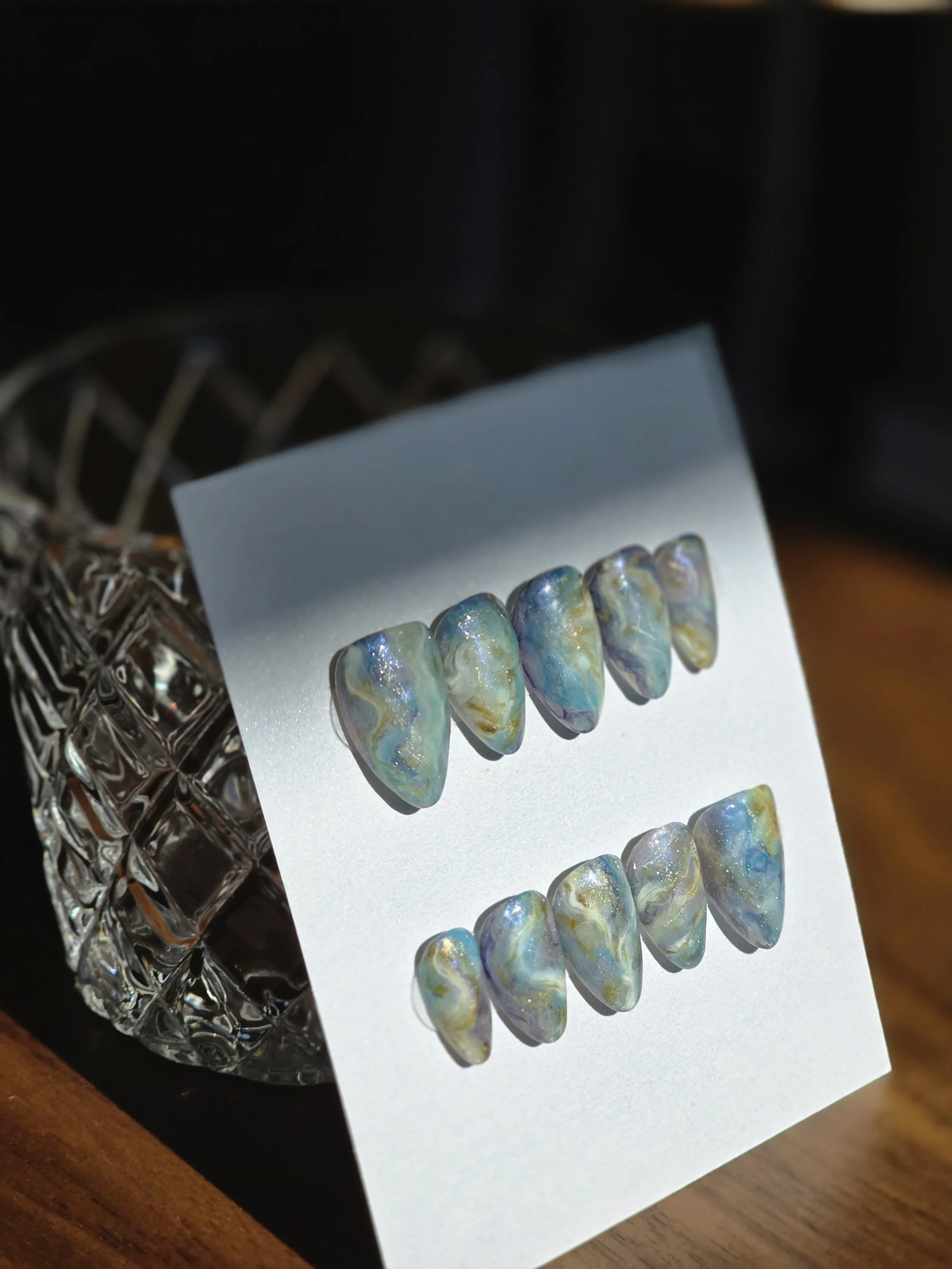 A display of iridescent, multicolored fake nails on a white card, with a silver crystal bowl in the background.