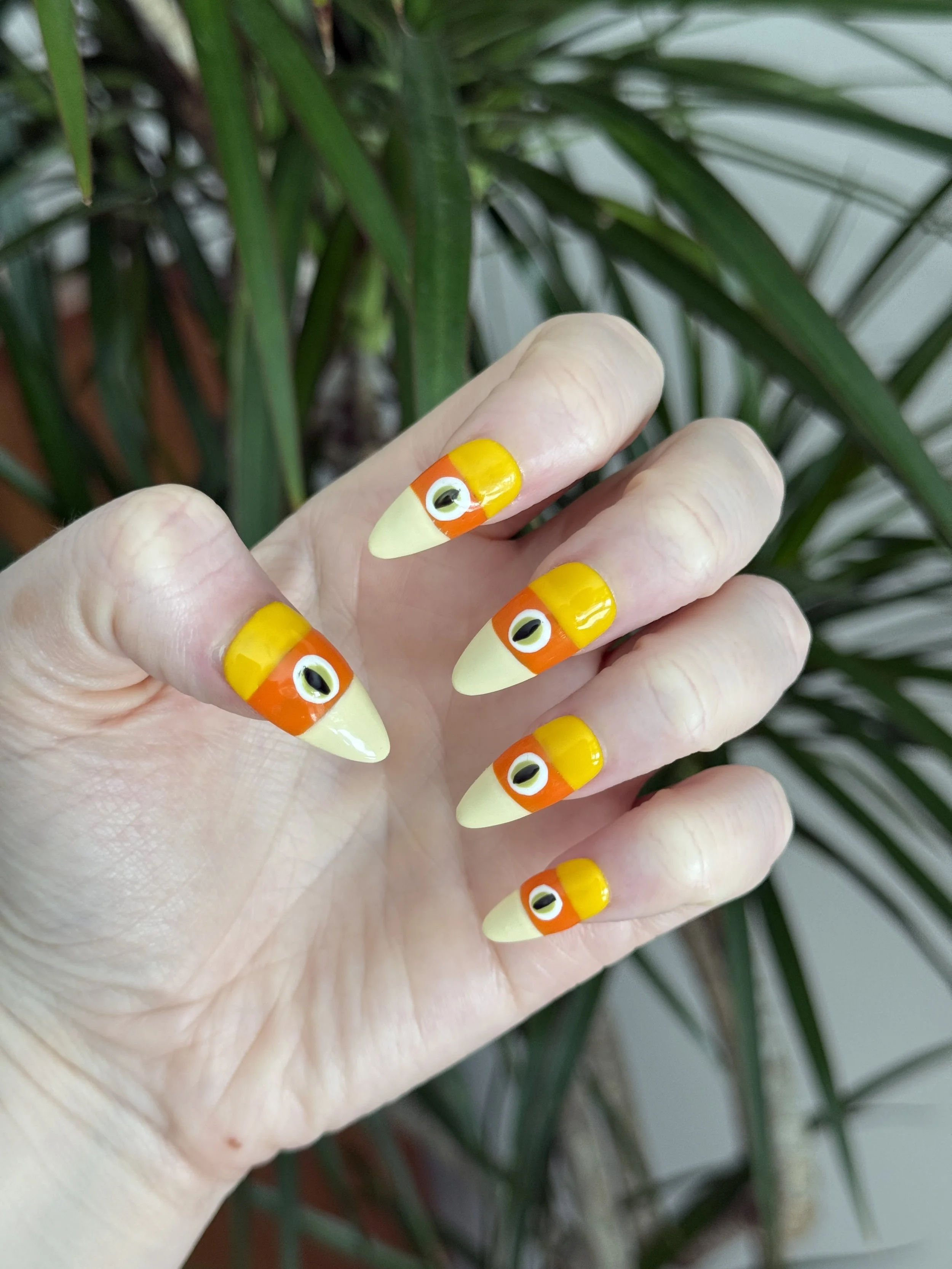Hand with cartoonish fish nail art, yellow and orange striped fingernails with black-and-white eye design, against green leafy background.
