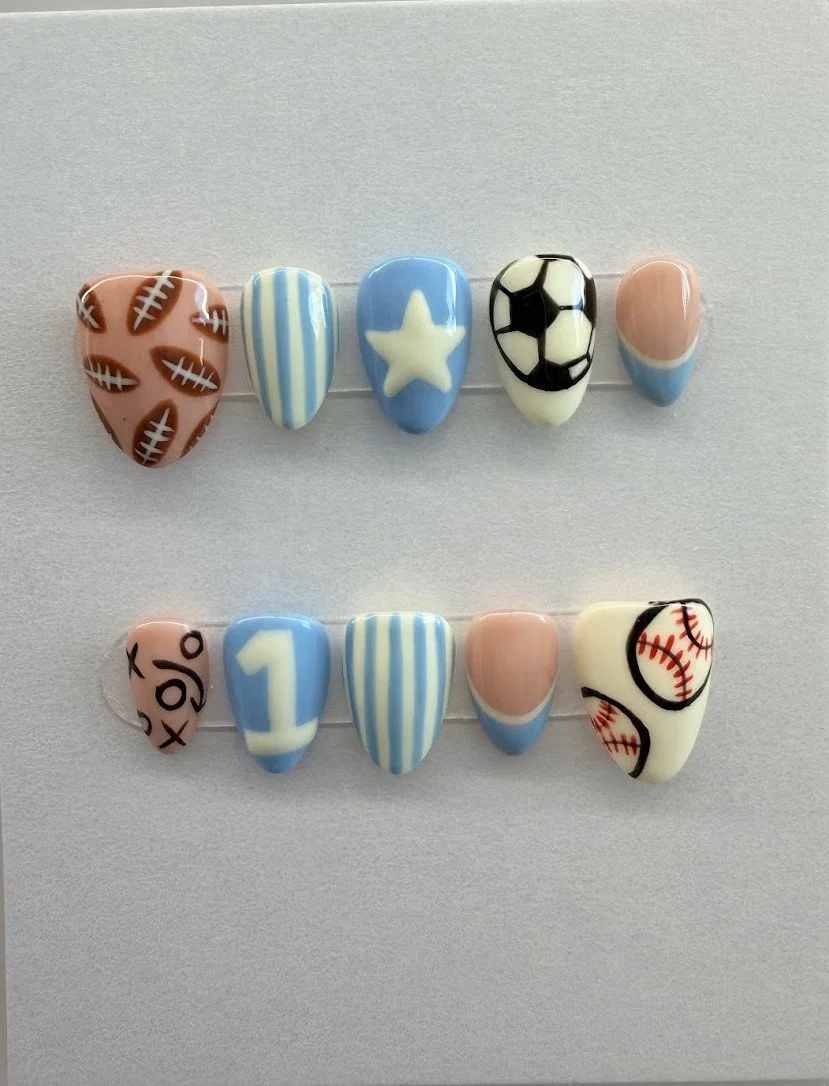 Two rows of decorated nail tips with sports theme designs. The top row features football, striped, star, soccer ball, and volleyball designs. The bottom row features a baseball, number one, striped, peach, and baseball pattern designs.