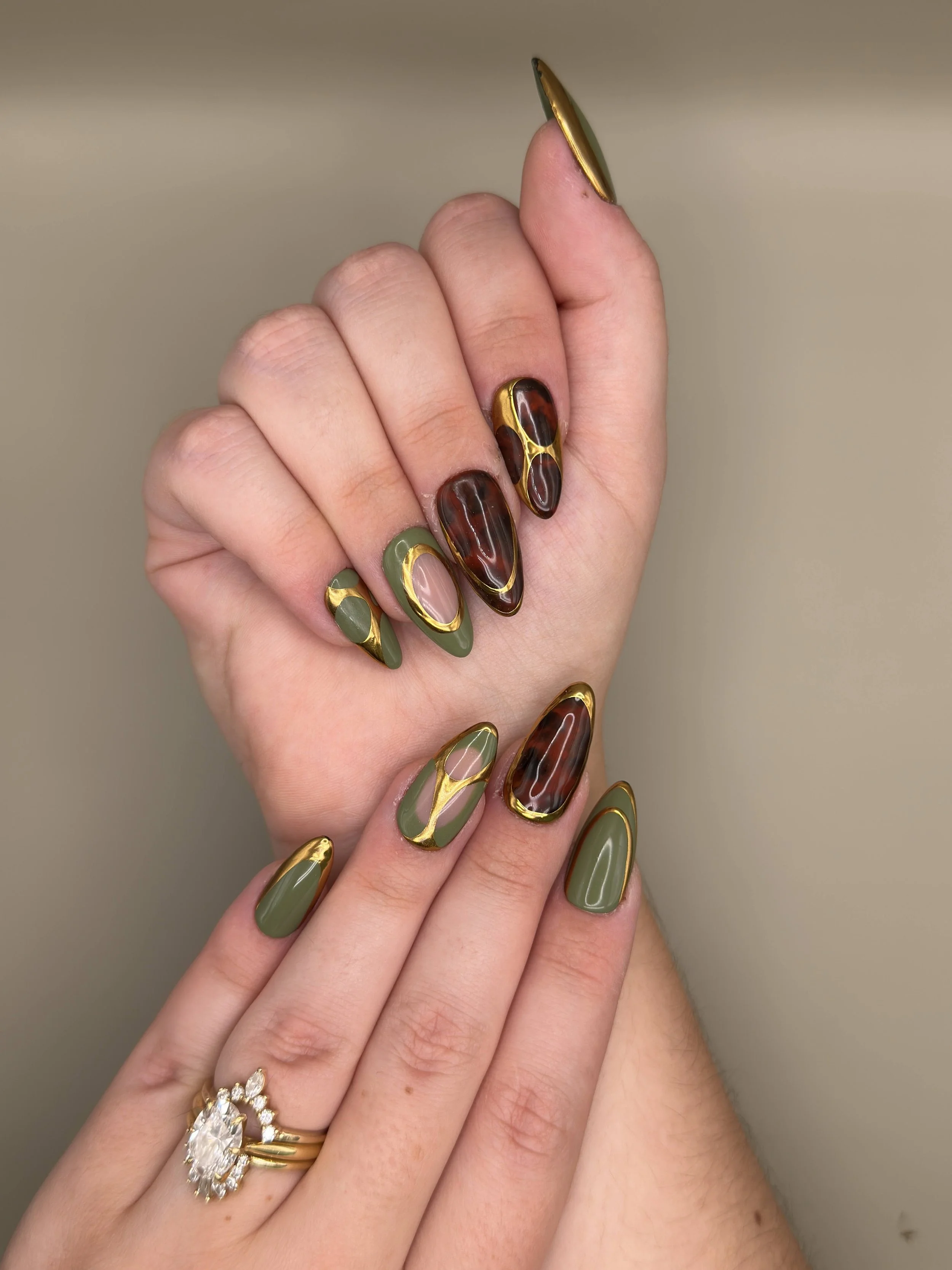 A woman's hand with elaborate nail art featuring tortoise shell and olive green designs, accented with gold details, and a gold ring with a large central gemstone surrounded by smaller stones.