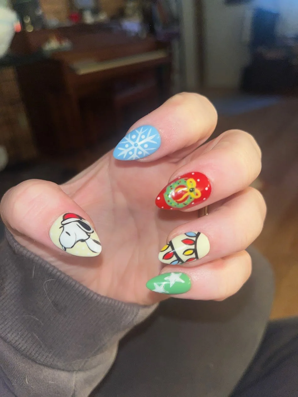 Hand with colorful Christmas-themed nail art, including a snowflake on the index finger, a Christmas ornament, a Snoopy dog, a Christmas light strand, a red ornament, and a green star on white and red bases.