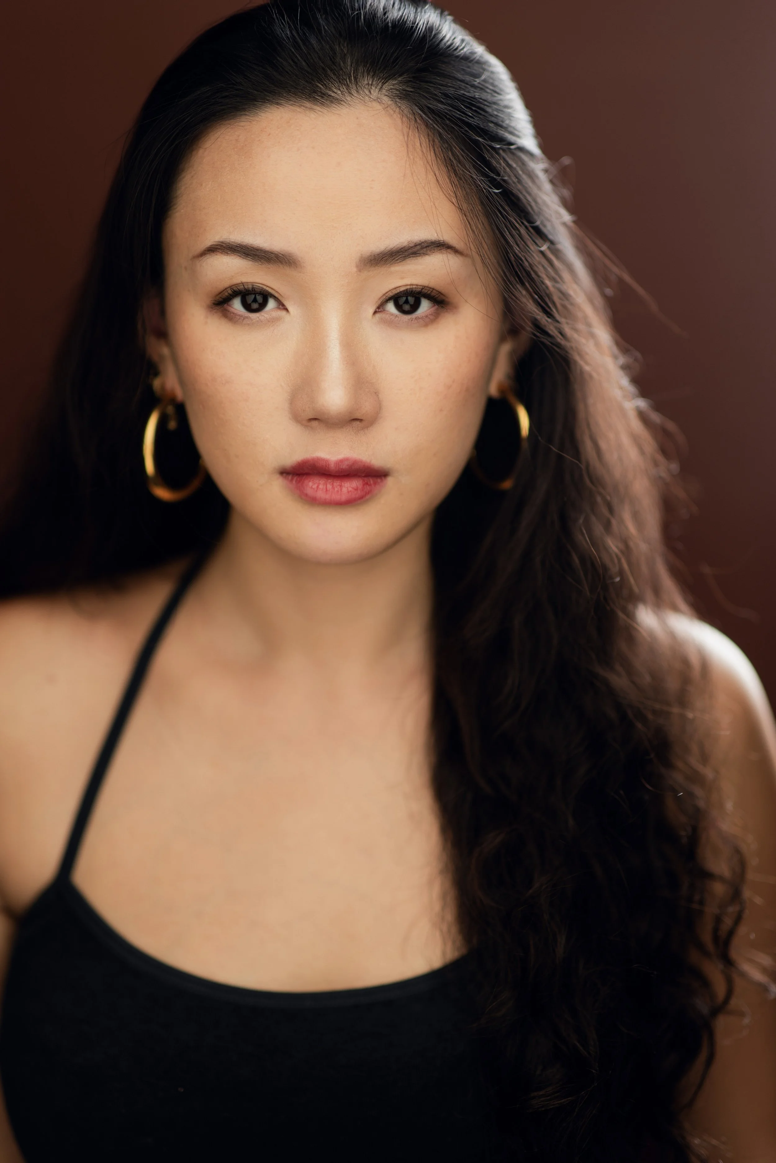 Close-up portrait of a young woman with long, dark wavy hair, wearing gold hoop earrings and a black top, with a neutral background.
