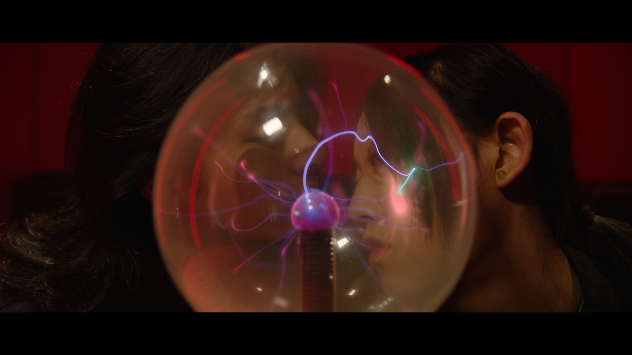 Two women with dark hair and earrings face each other closely, observing a plasma ball with colorful electrical discharges inside, set against a red background.