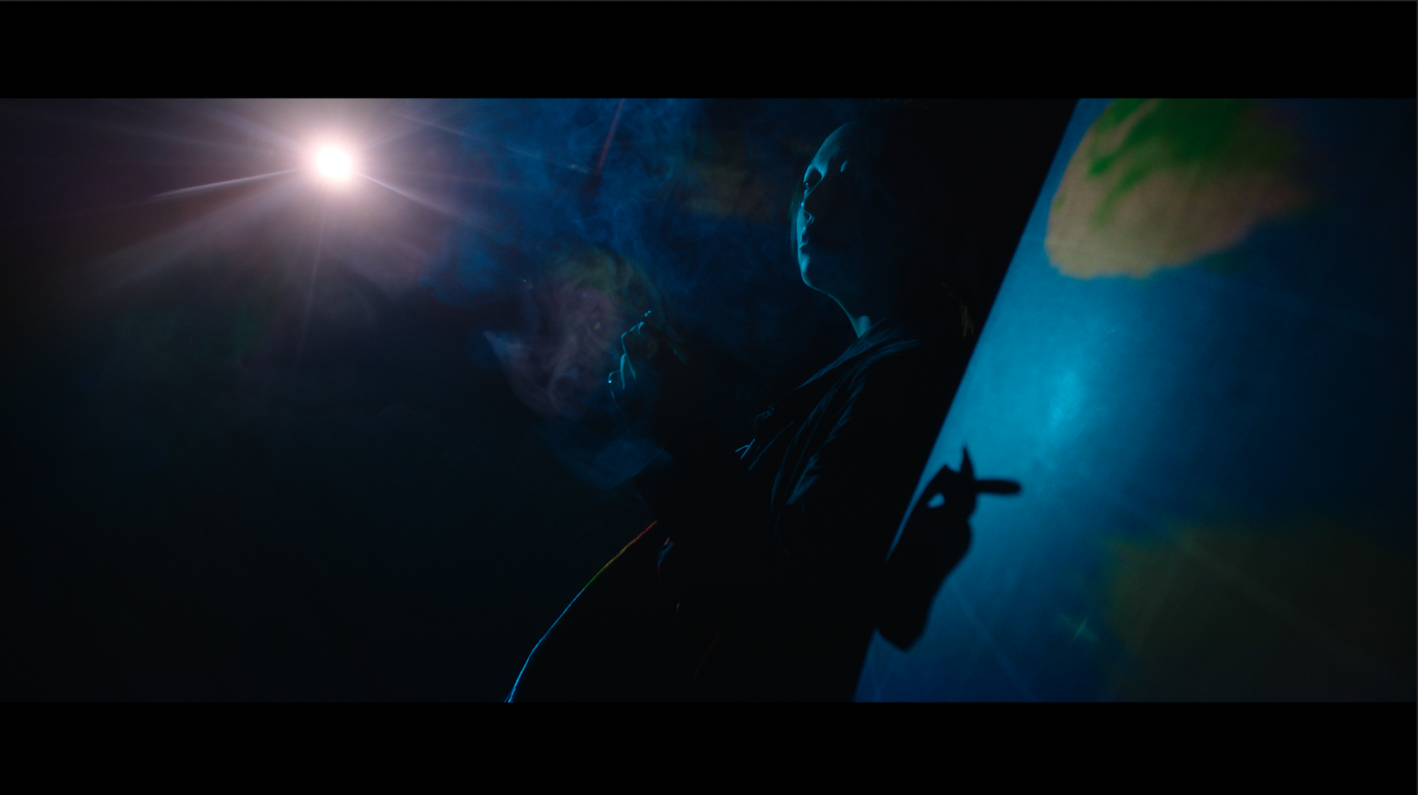 A person sitting next to a world map, smoking and illuminated by an overhead light in a dark room.