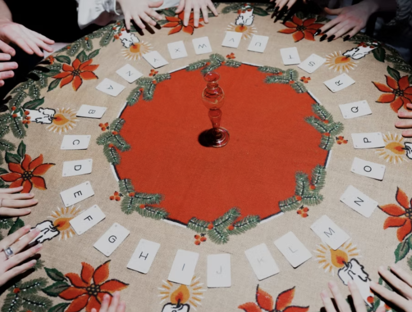 Children's hands surrounding a Christmas-themed table with a red and green wreath, alphabet cards, and a candle centerpiece, engaged in a word or alphabet game.