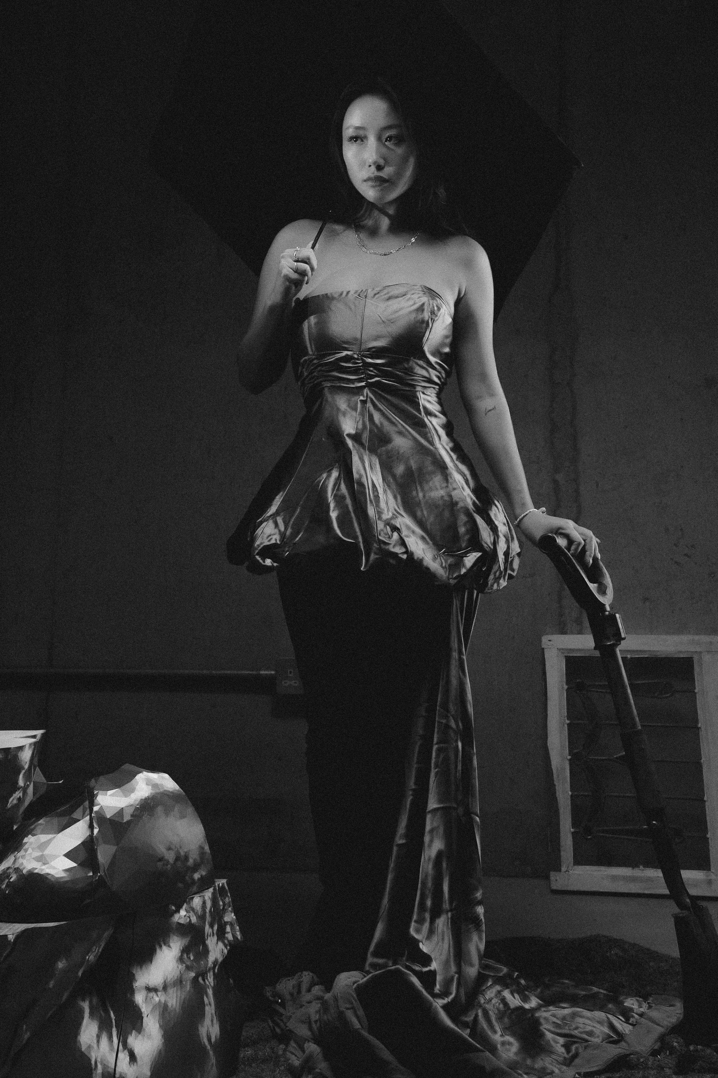 A woman standing with an umbrella, wearing a shiny strapless top and a long skirt, holding a bag in her left hand, in a dark room with a rough wall and a small window.