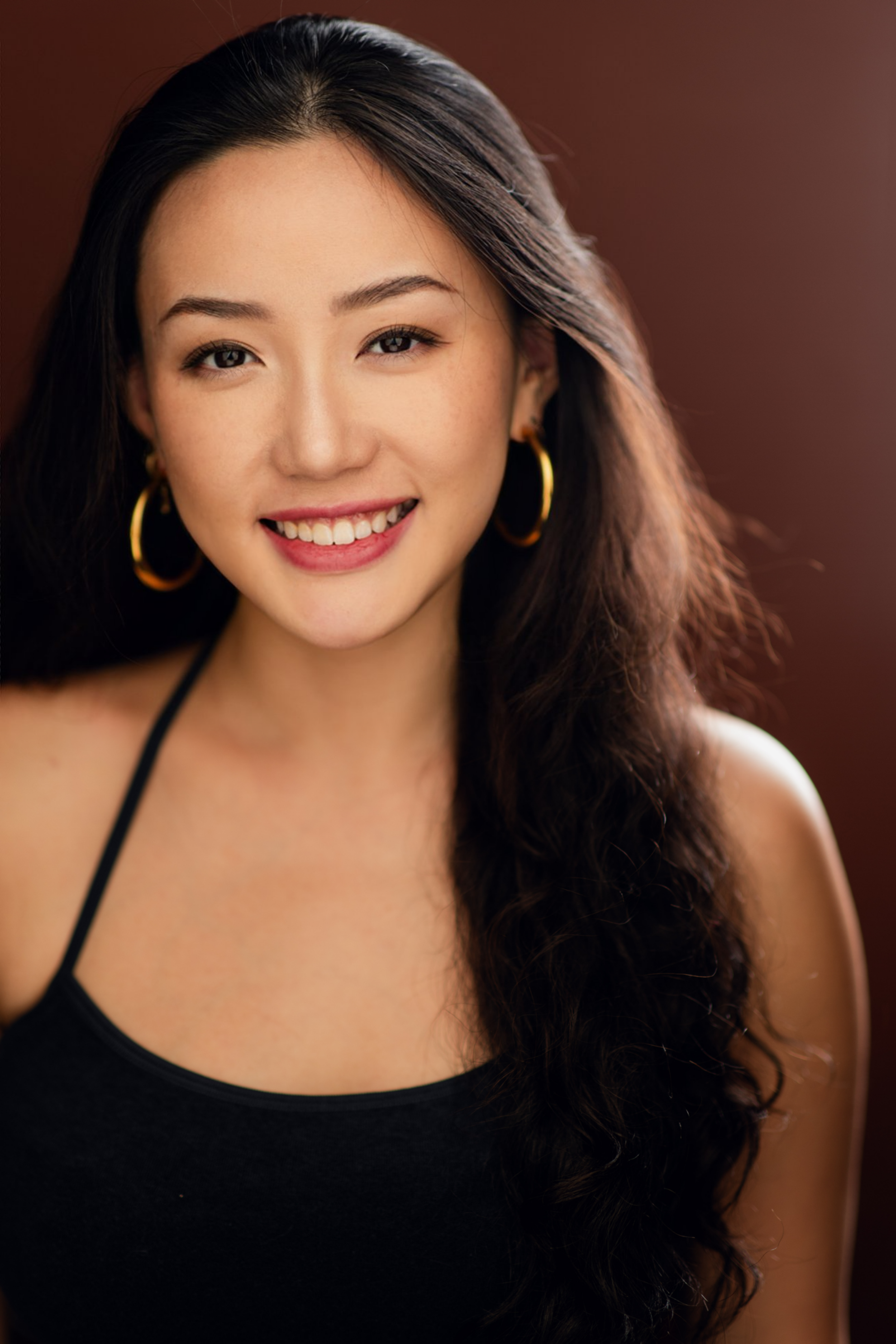 A smiling young woman with long dark hair wearing gold hoop earrings and a black top.
