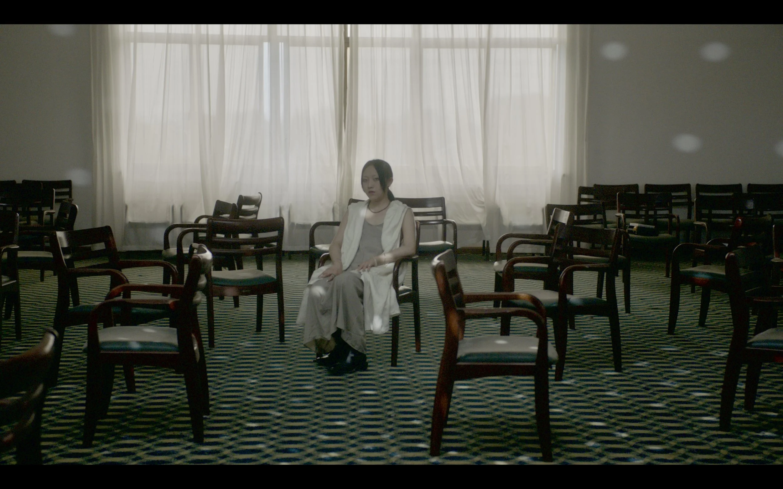 A woman sitting alone in an empty room with numerous wooden chairs, closed light-colored curtains, and patterned carpet.