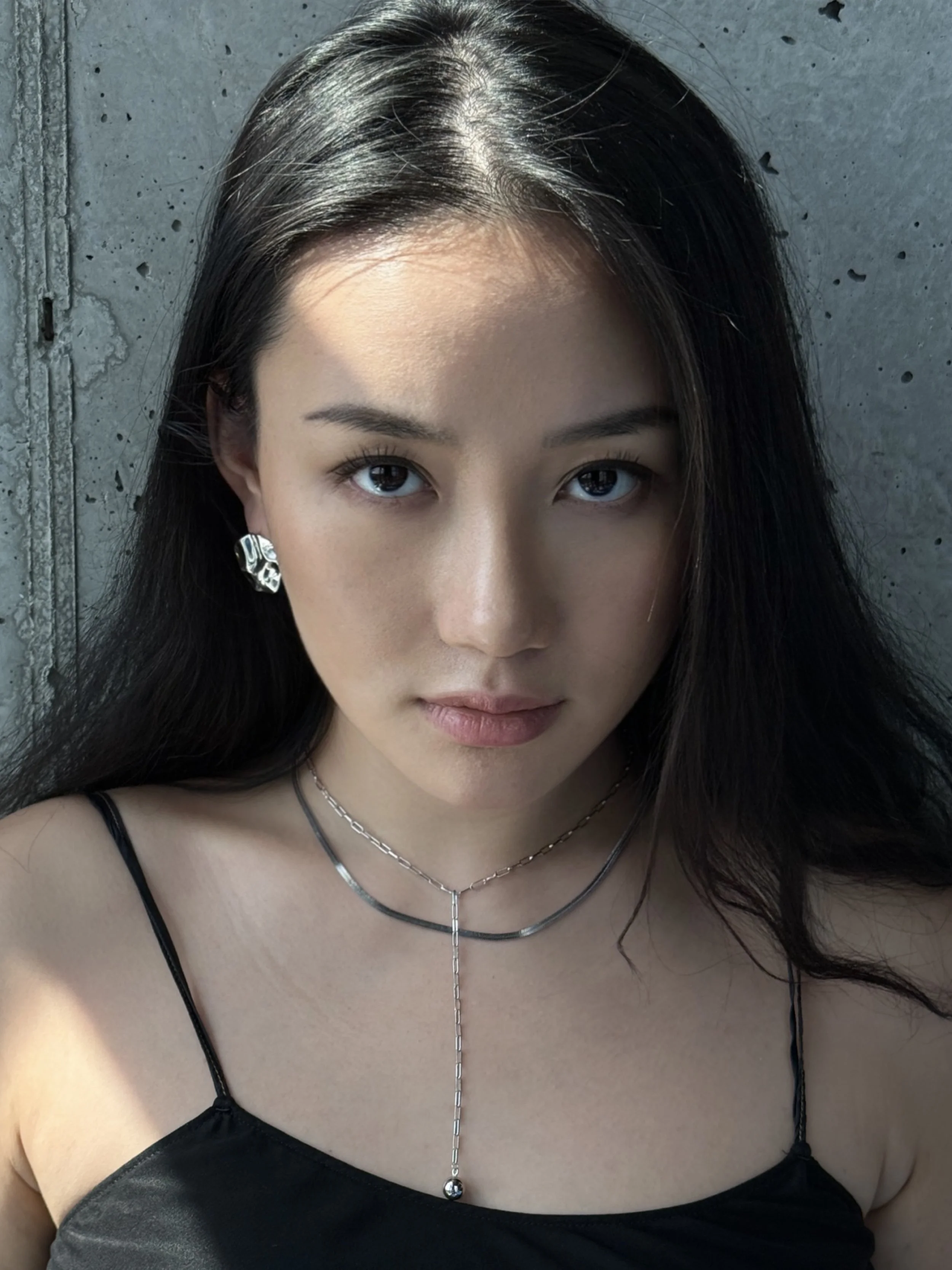 A young woman with long dark hair, wearing earrings and layered necklaces, looking directly at the camera with a neutral expression against a concrete wall background.