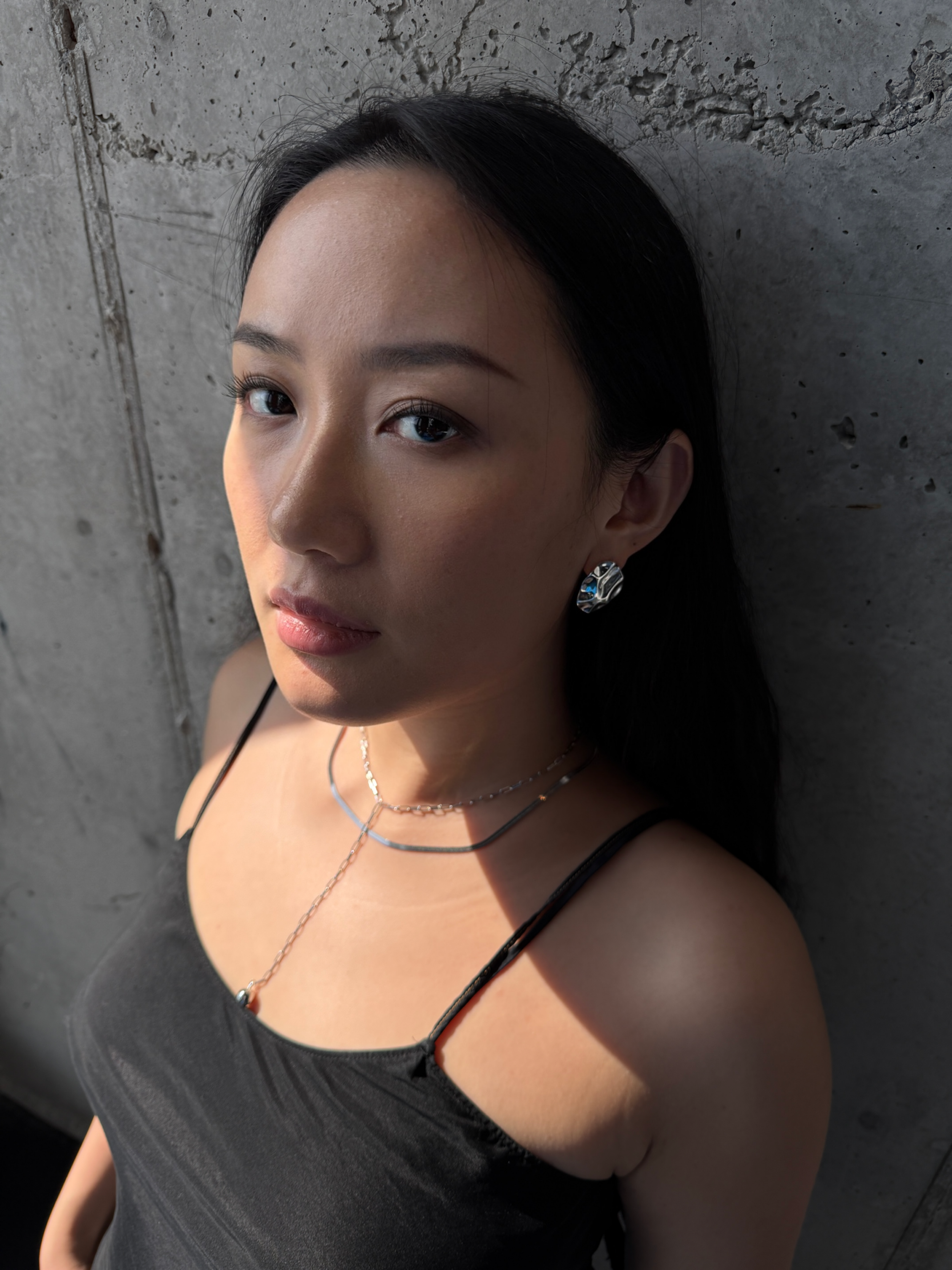 A woman with dark hair and makeup, wearing a black spaghetti strap top, silver jewelry, and standing against a concrete wall.
