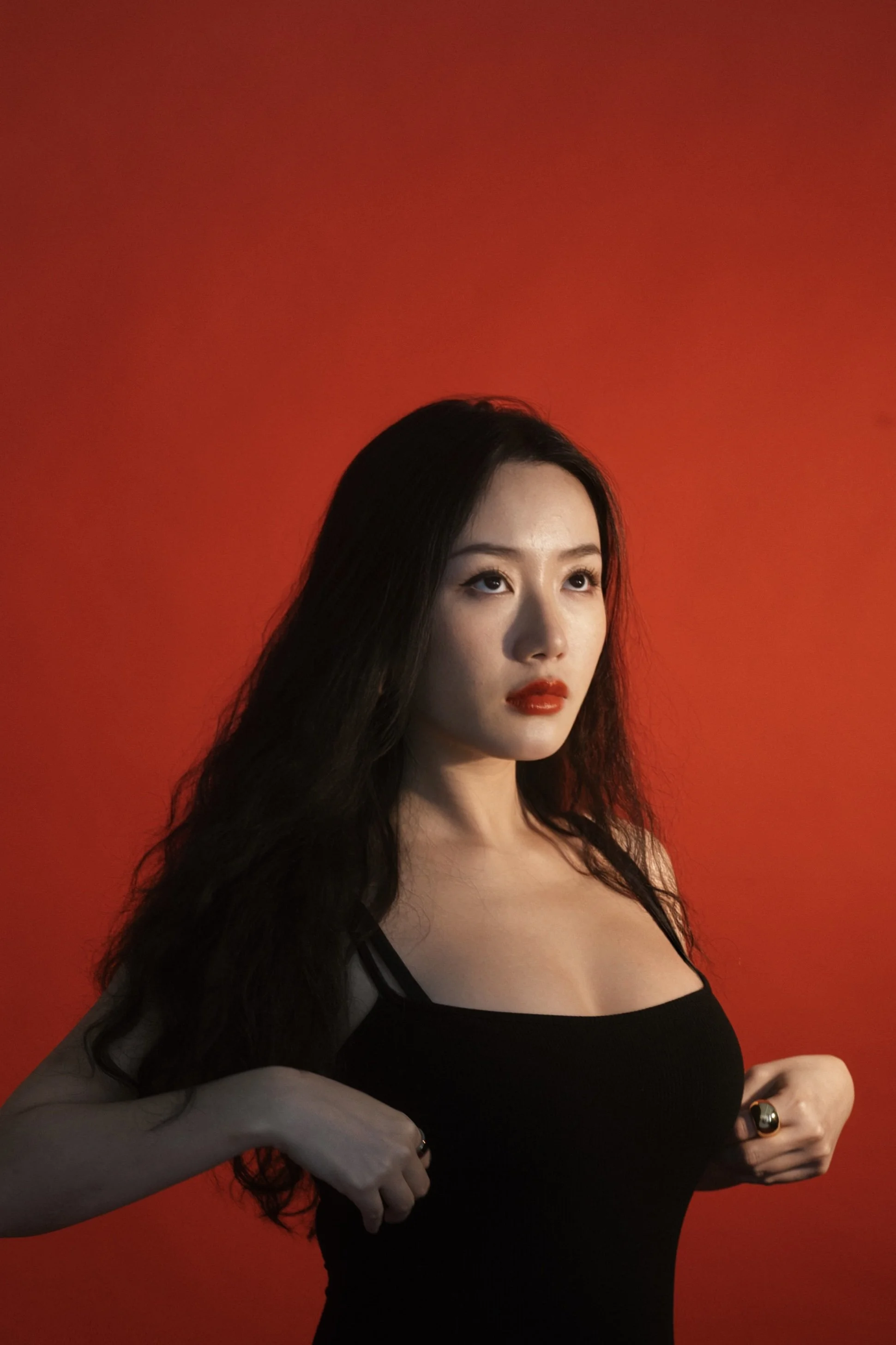 A woman with long black hair, wearing a black top, stands against a red background. She has red lipstick and is holding her fists near her waist.