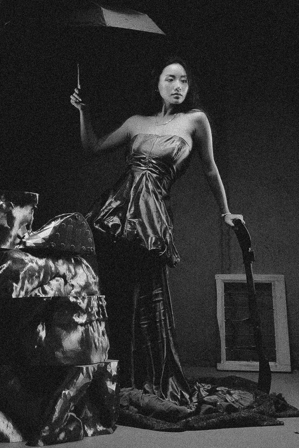 A woman in a strapless dress holding an umbrella stands next to a patterned object, with an open door in the background.