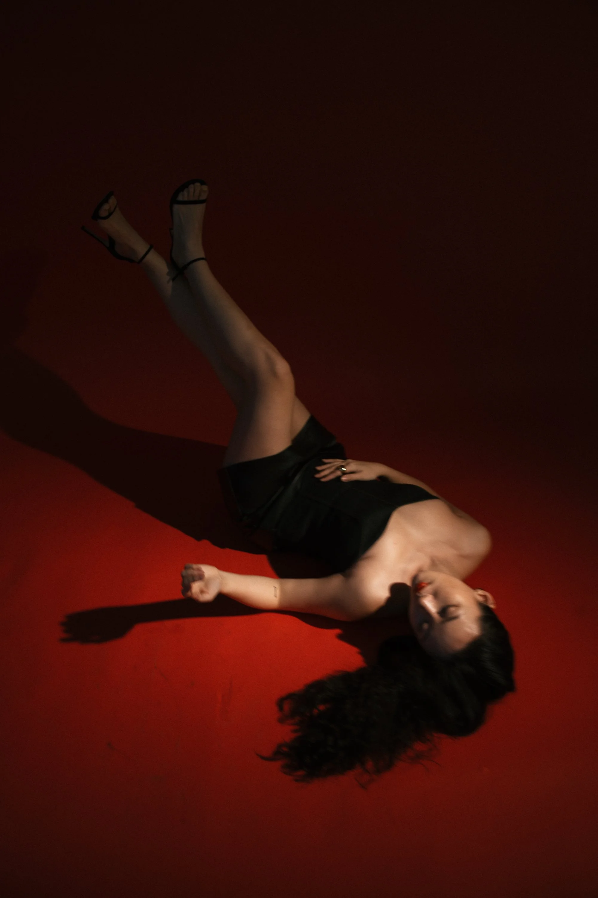 A woman with long, curly dark hair lying on a red floor in a black dress and high heels, casting a shadow. She is positioned on her side with eyes closed.