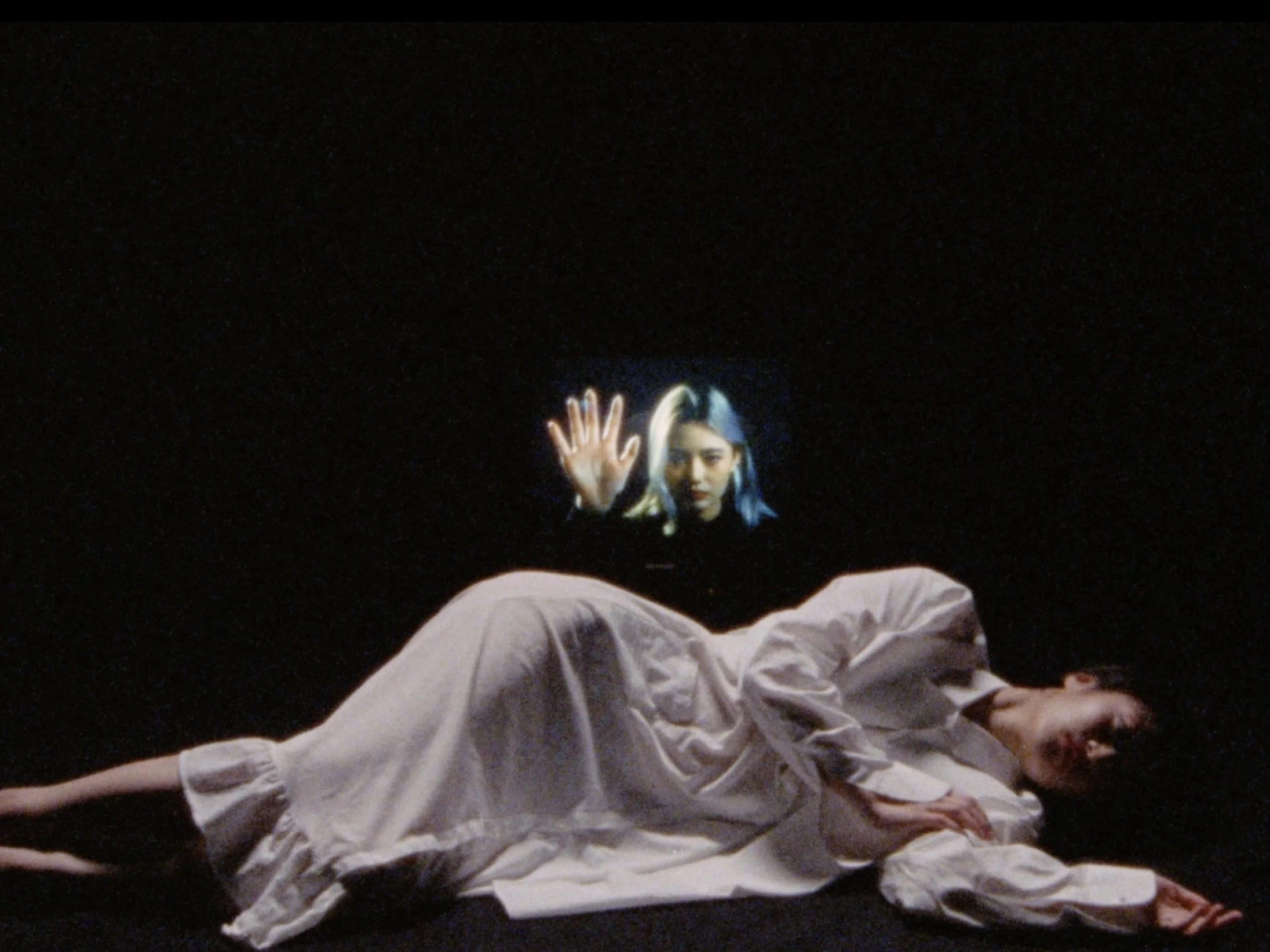 A woman laying on the floor in a white dress, with a focus on her face and body. A virtual holographic projection of another woman with dark hair and a serious expression appears above her, waving her hand.