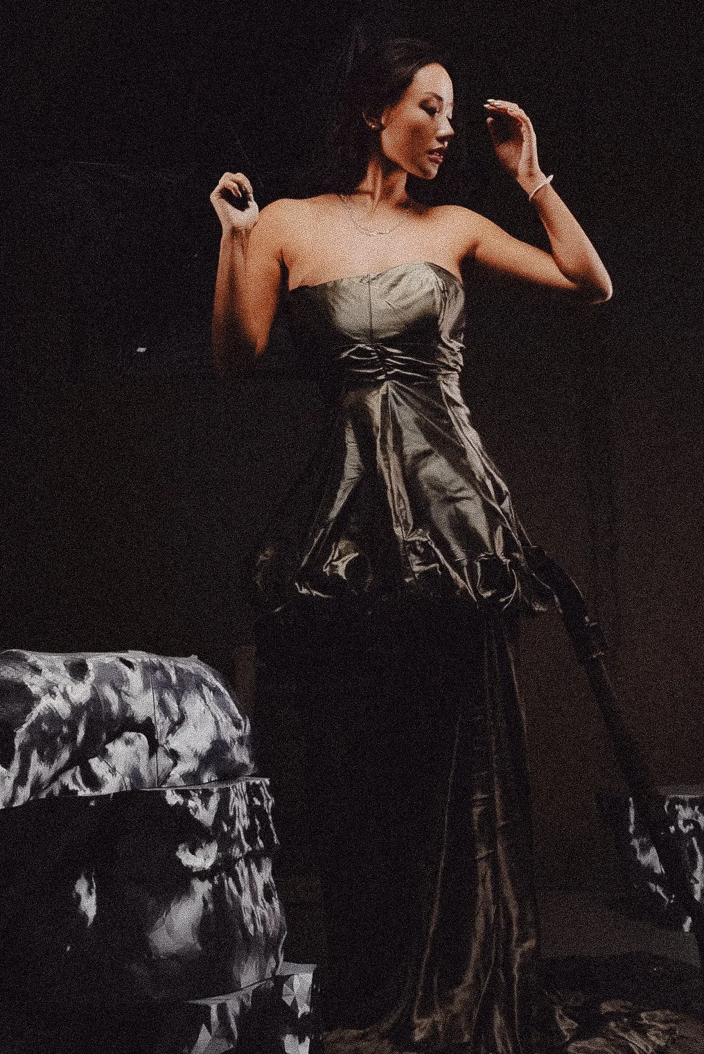 A woman in a strapless metallic dress is posing against a black background, with her arms raised and eyes closed.