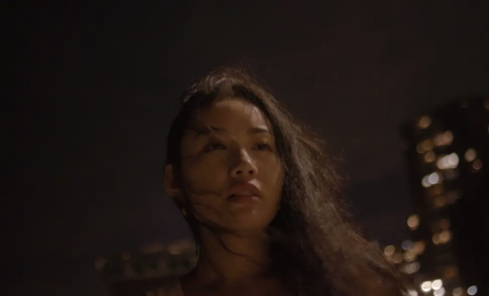 A woman with wind-blown hair at night against a cityscape with blurred city lights.