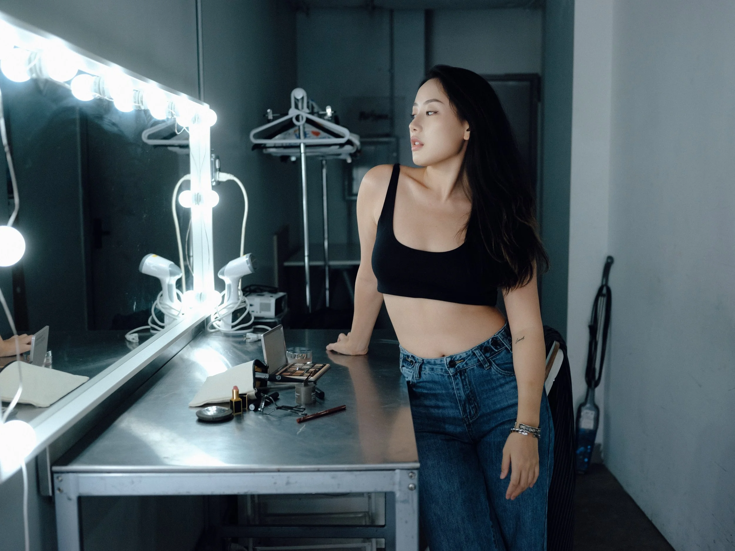 A young woman with long dark hair, wearing a black crop top and blue jeans, stands in front of a mirror with bright lights. She is in a dressing room or backstage area, with makeup and beauty products on the counter.