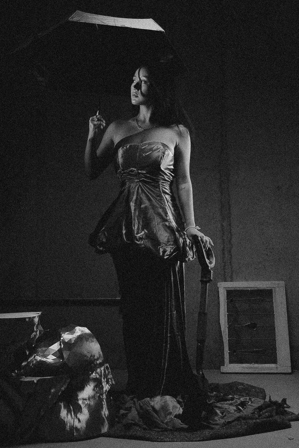 A black and white photograph of a woman standing with an umbrella in a dark room. She is dressed in a strapless gown, holding her right hand up, and has a contemplative expression.
