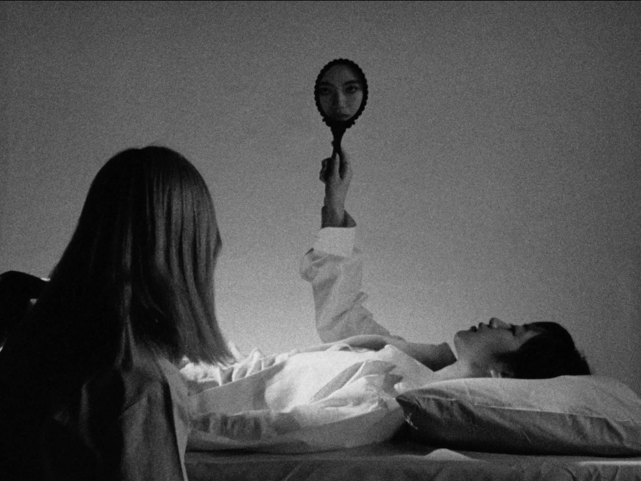 A woman lying in bed looking up, another woman sitting beside her, and a mirror held above the bed showing a close-up of her face.