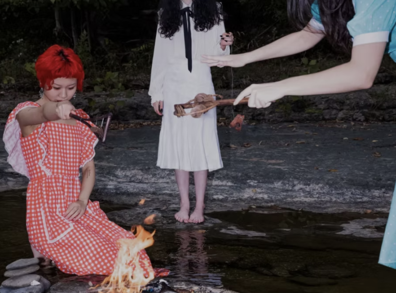 Three people around a small fire near a river, with one person roasting a marshmallow, another holding tongs, and the third standing barefoot.