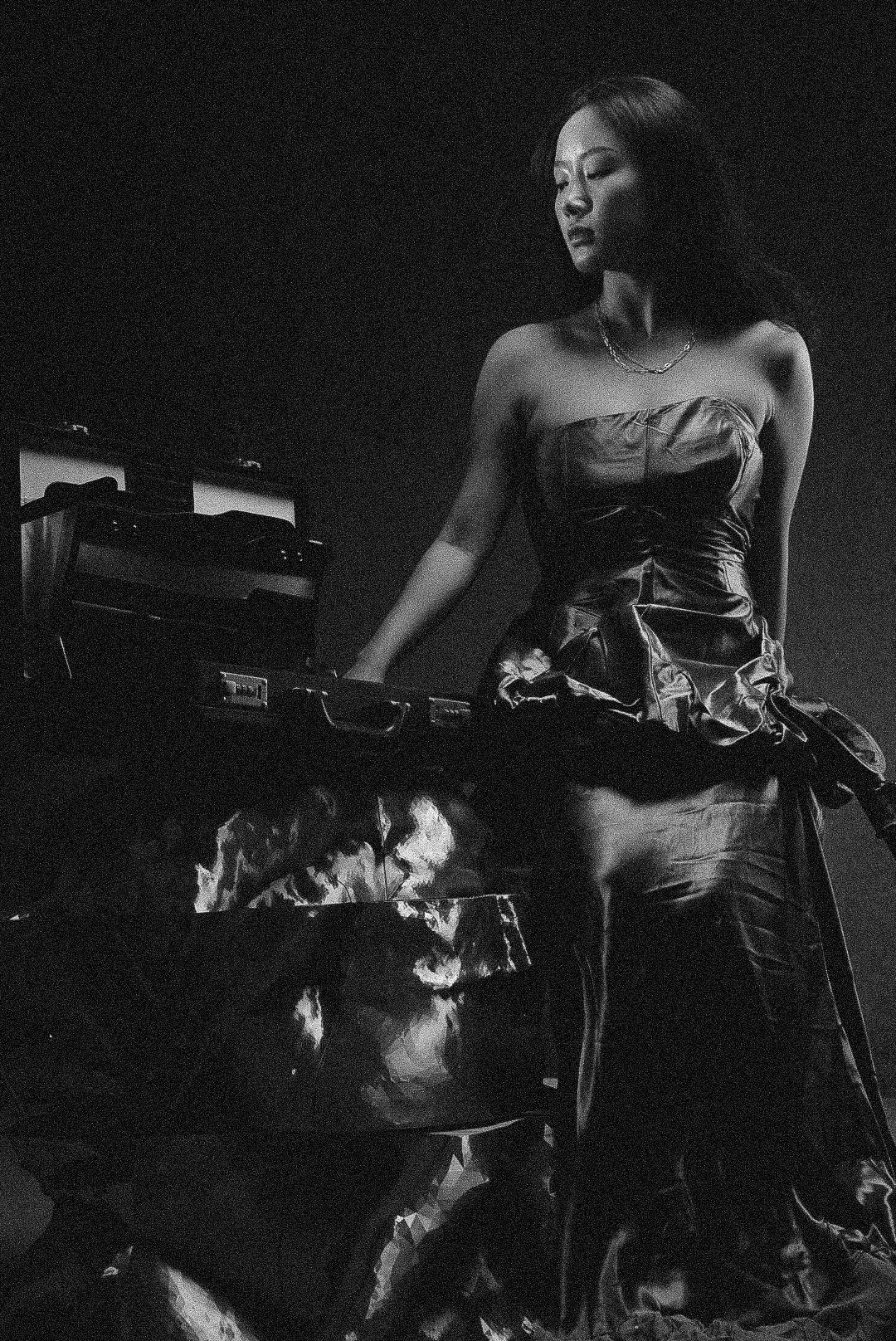 A woman with long hair standing next to a piano, dressed in a strapless shiny dress, in a dark room with minimal lighting.