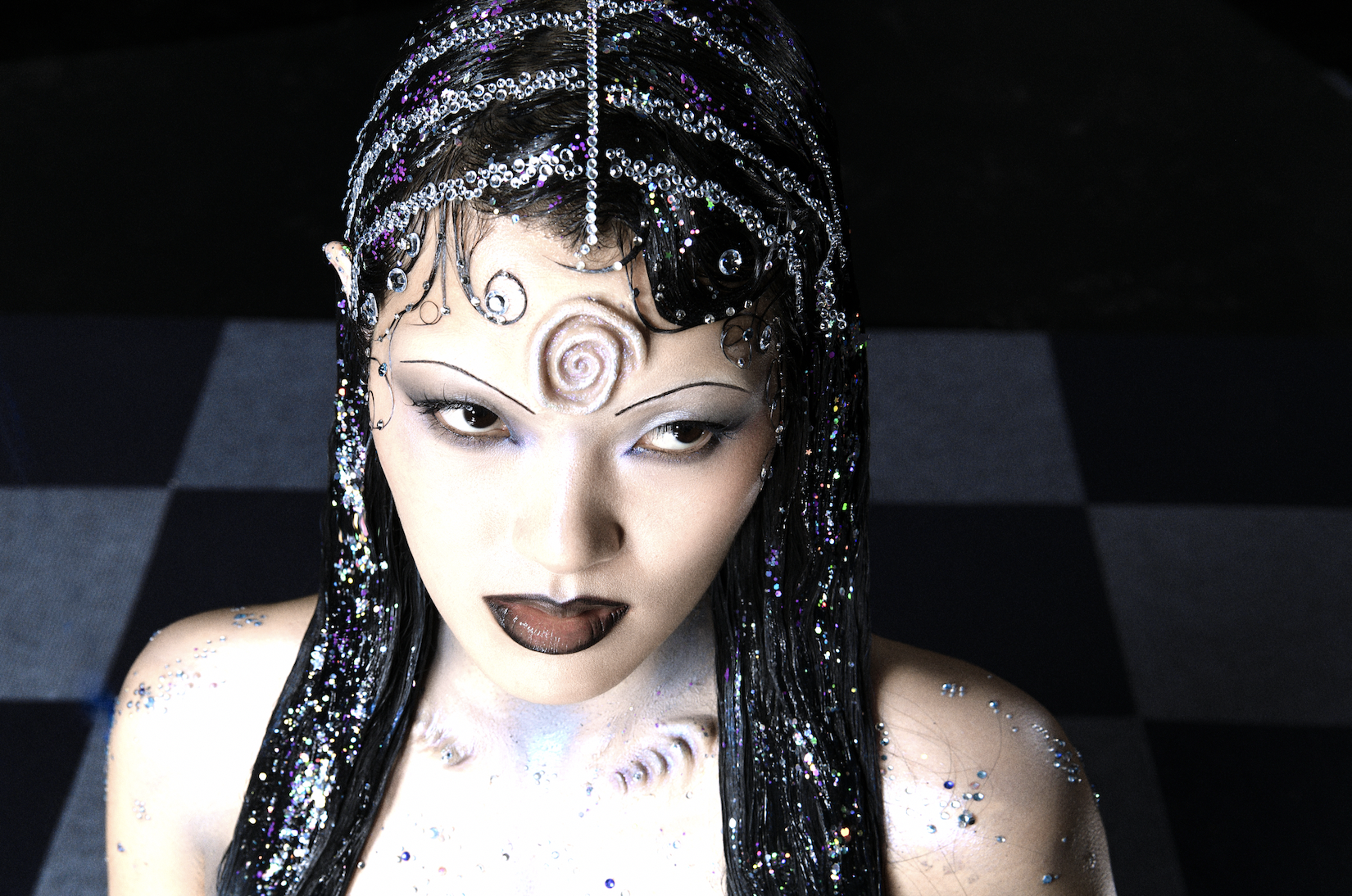 A woman with elaborate black hair styled with silver and purple beads, adorned with sparkly accessories, has face and body makeup with white and black tones, and a spiral design on her forehead. She wears dark lipstick and dramatic eye makeup, and has rhinestones on her shoulders.