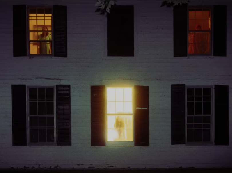 A two-story house with six windows, three on each floor. The middle window on the lower floor is bright, with a human figure visible through the glass. The upper windows have glowing light and curtains, with two showing silhouettes of people.