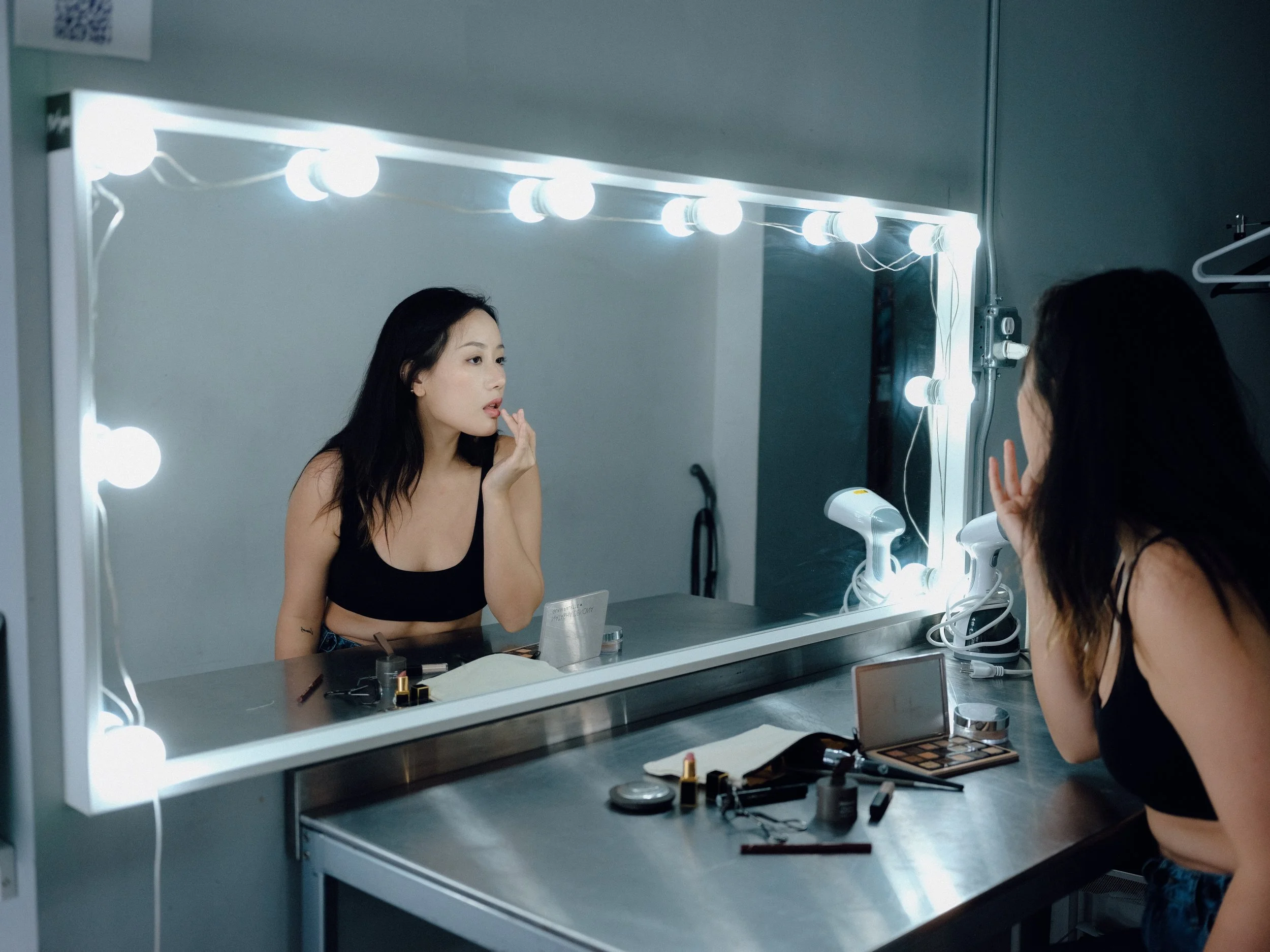 Woman applying makeup at a vanity mirror with bright lights, makeup products and tools on the table.