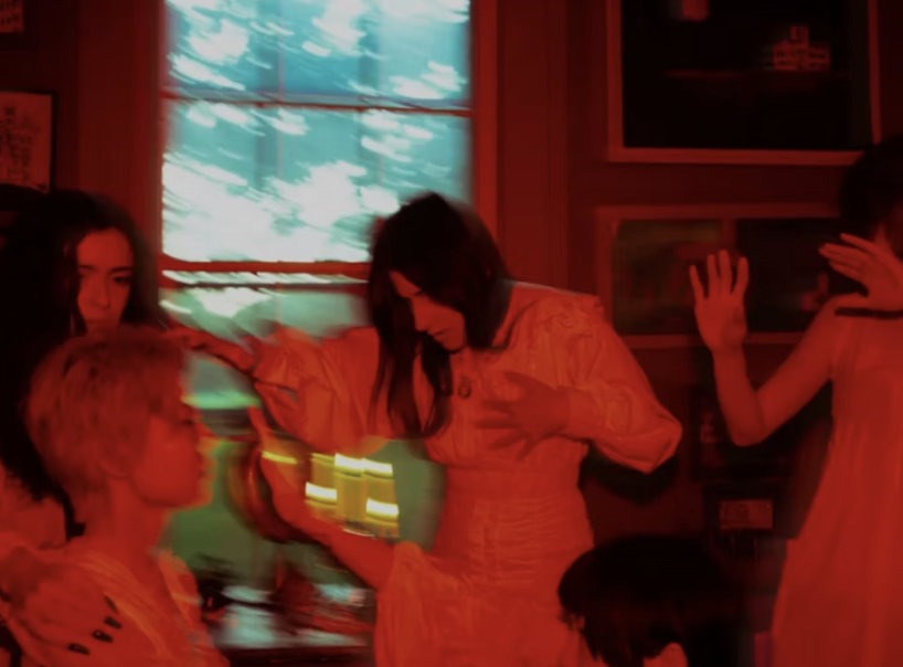 Four young women in a room with red lighting, some with their hands raised and one leaning against a window.
