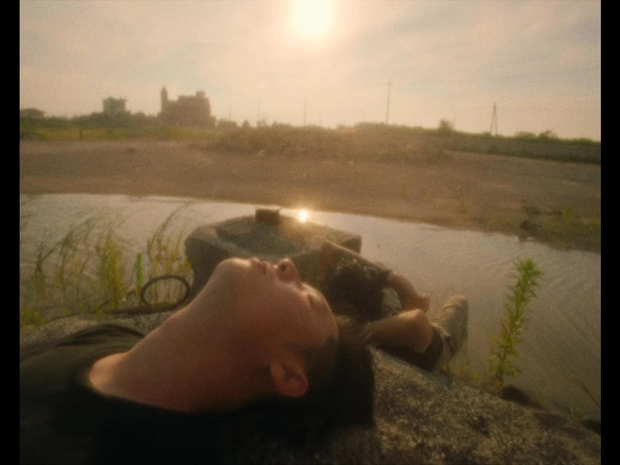 A person lying down outdoors near a body of water, with their face turned upward and eyes closed, during sunset.