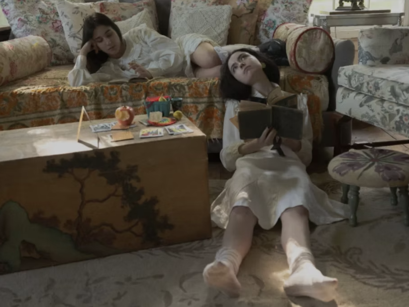 Two women relaxing in a cozy living room, one lying on a floral-patterned sofa, and the other sitting on the floor reading a book, with a wooden coffee table and floral upholstery nearby.