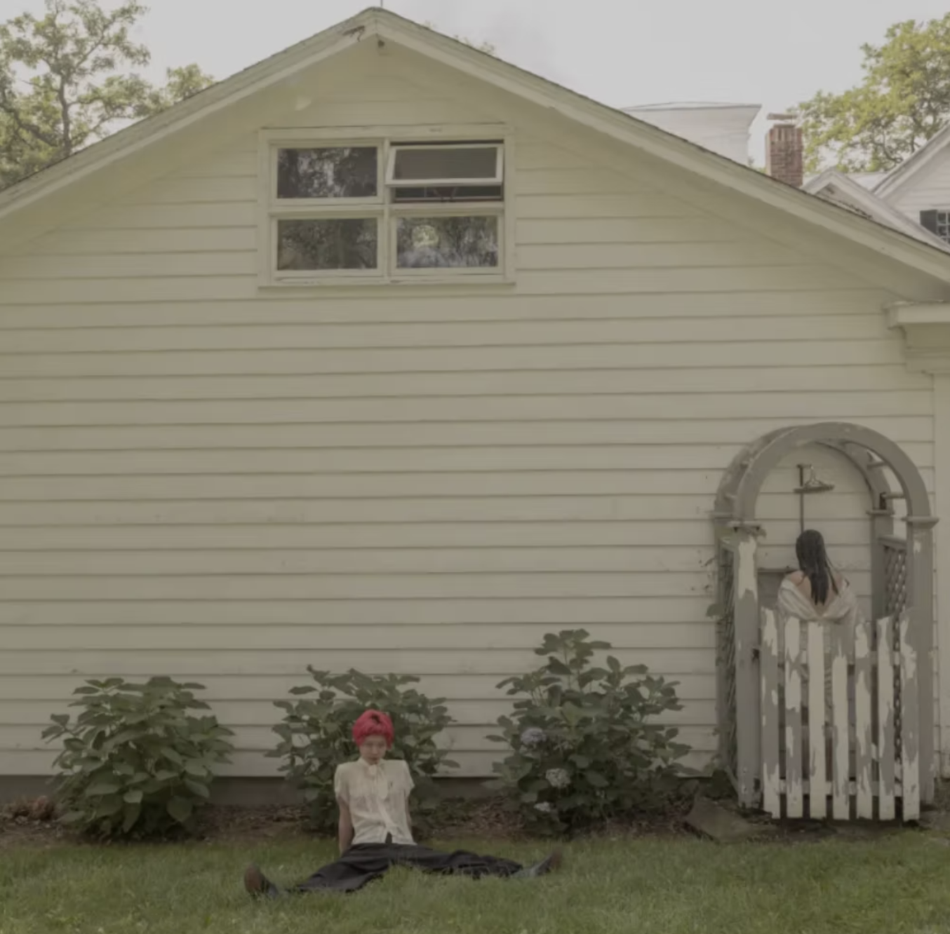 A white house with horizontal siding, two windows, and a small yard with green plants. A woman with pink hair sits on the grass, and a person with long dark hair leans over a white fence archway.